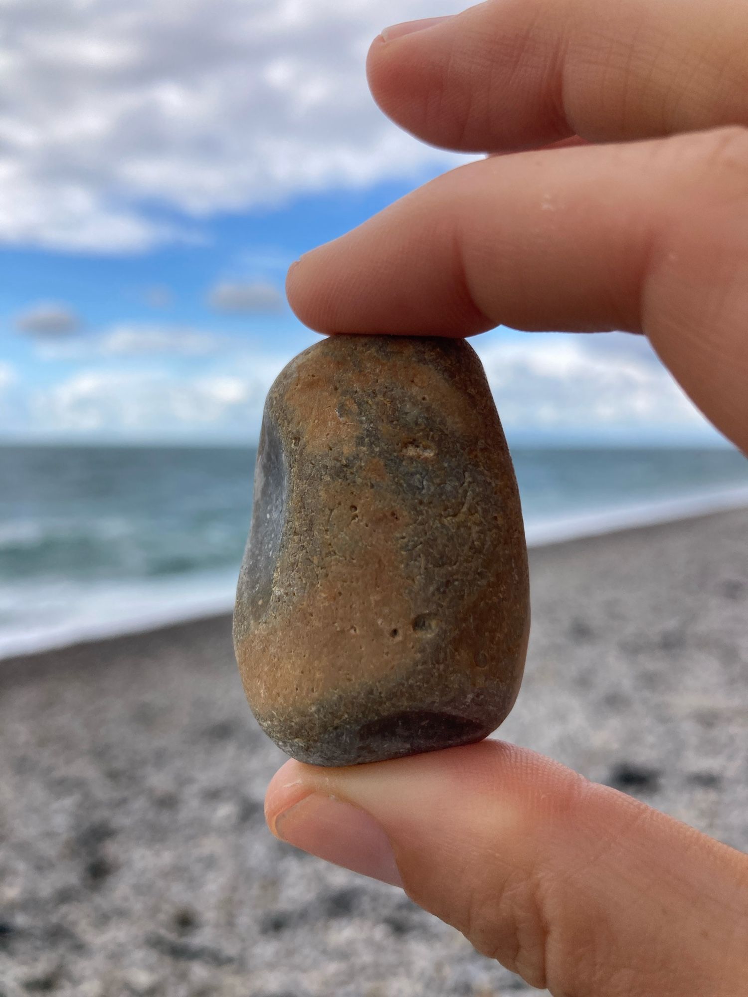 A hand holds a vaguely rectangular, black and brown beach pebble, mostly dull but with a shiny chip on one side and a smaller one near the bottom. The backdrop is a rocky beach, the sea, the sky.