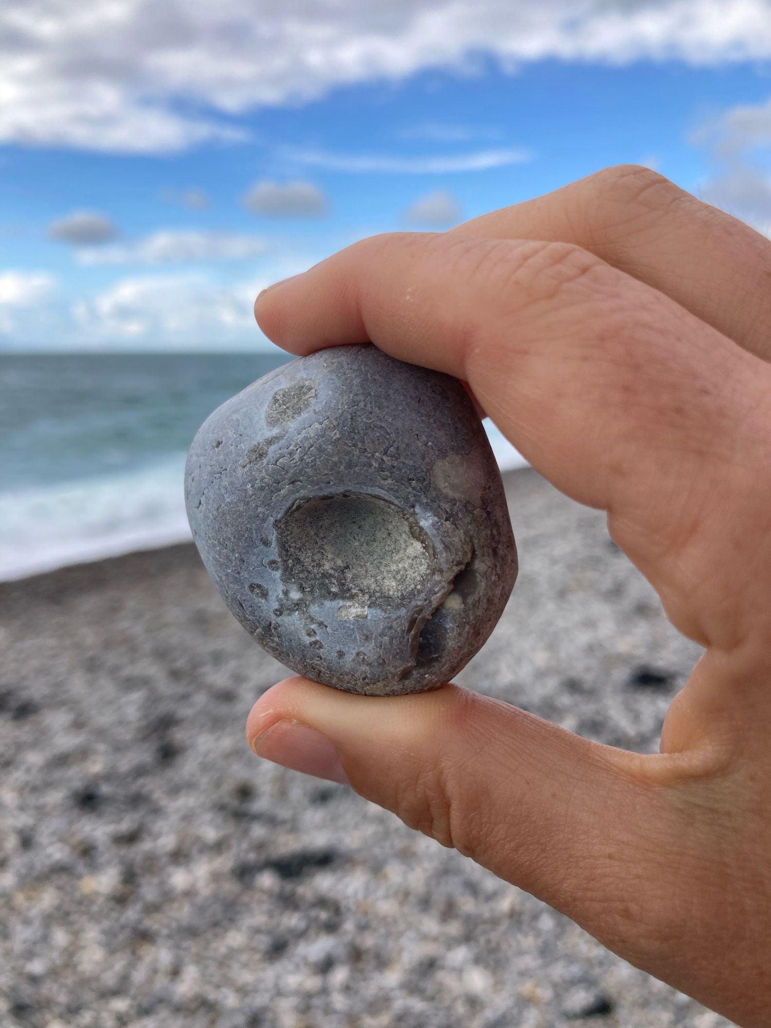 A hand holds a rounded grey beach pebble with a large hole near the middle, a smaller, bean-shaped one next to it, both brownish grey, and a couple of brownish round spots (one dark and one lighter). The backdrop is a rocky beach, the sea, the sky.