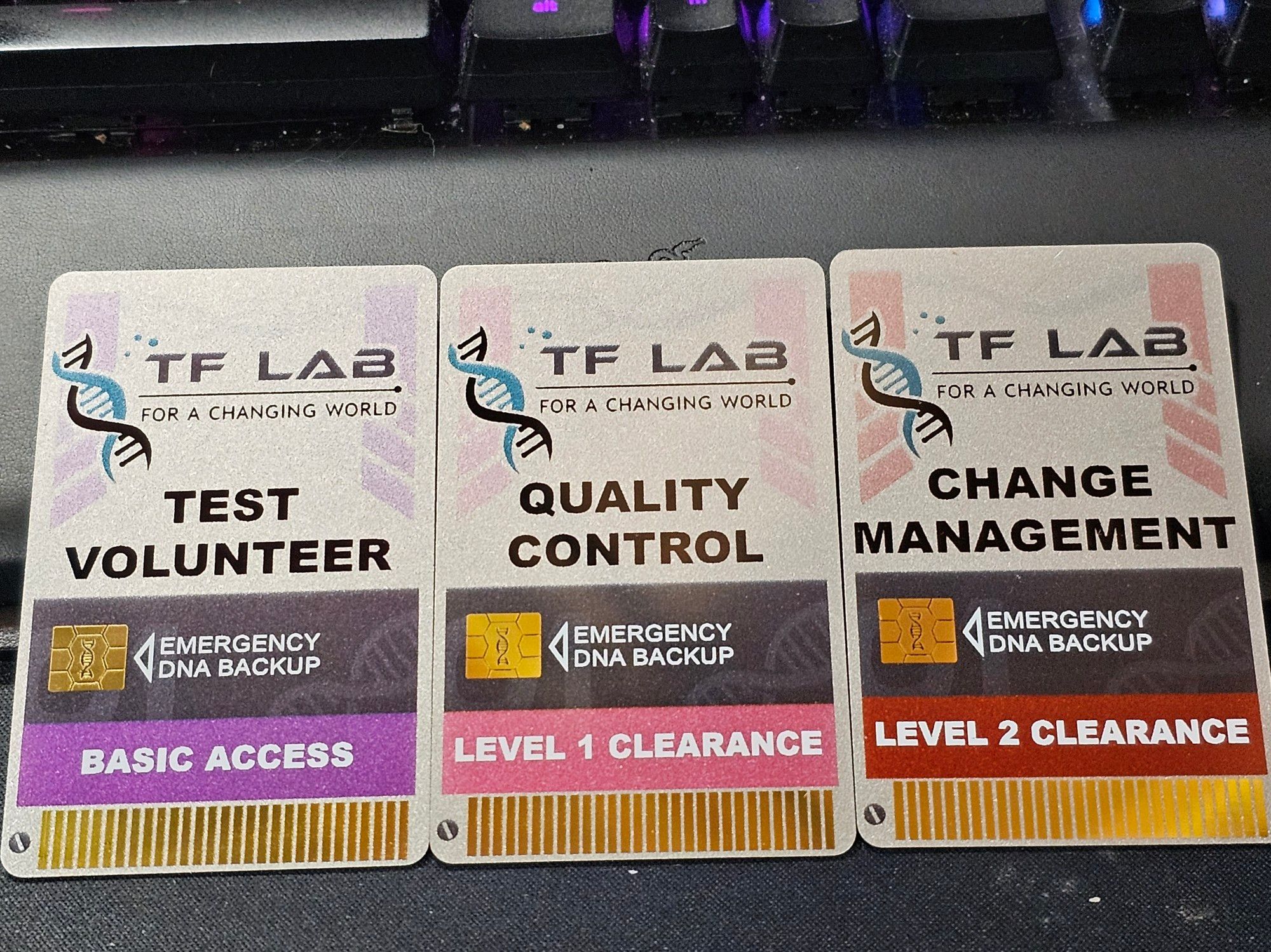 A photograph of 3 ID cards.

"TF Lab: for a changing world" is on all 3 of them, including a sim card spot for "emergency DNA backup"

The first card is "Test Volunteer" with basic access. The second is "Quality Control" with level 1 clearance. And the third is "change management" with level 2 clearance