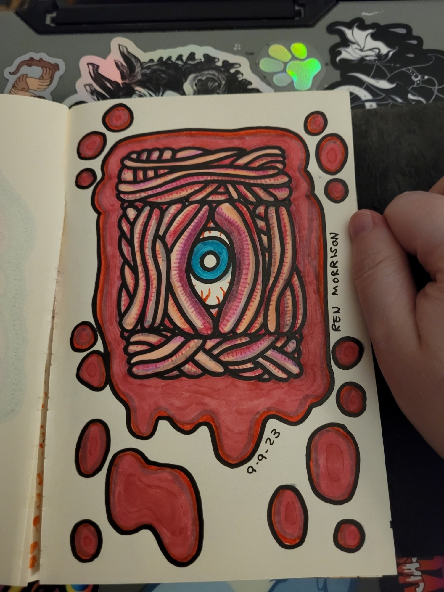 Photo of marker drawing art of an eyeball in a rectangle of flesh colored wormy looking things. Eye has a blue iris. The drawing is surrounded by a frame of splattered red marker. Art by Ren Morrison