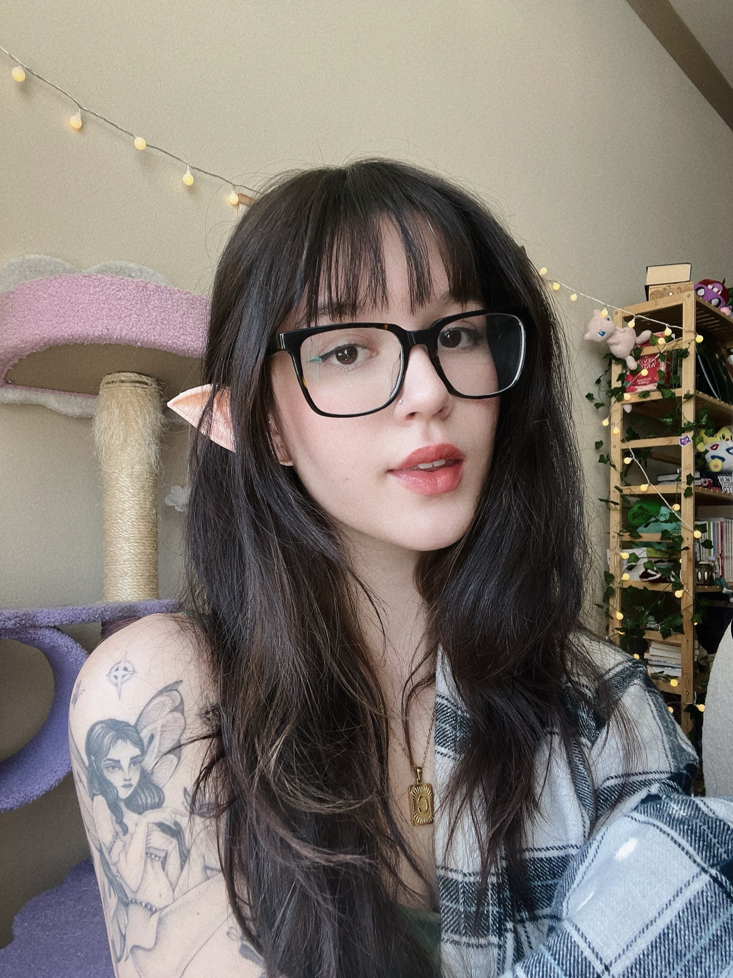 A selfie of Cass! They have long dark brown hair with bangs. Dark brown square glasses. A white and grey flannel hanging off her left shoulder, showing off her faerie tattoo. 