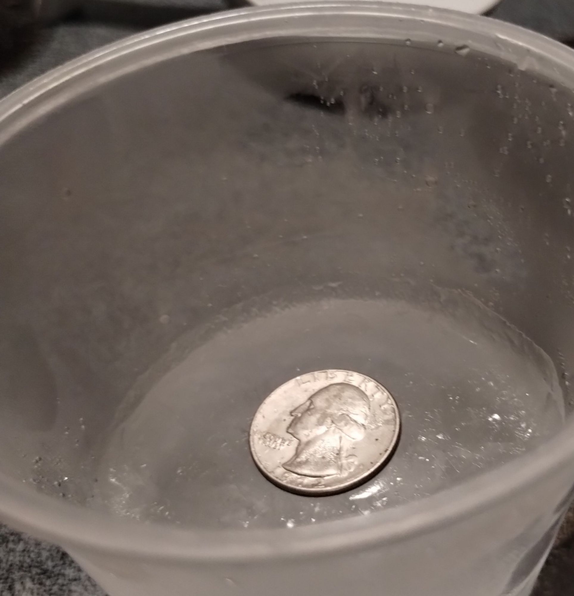 A plastic cup half full of frozen water with a quarter sitting on top of the ice.