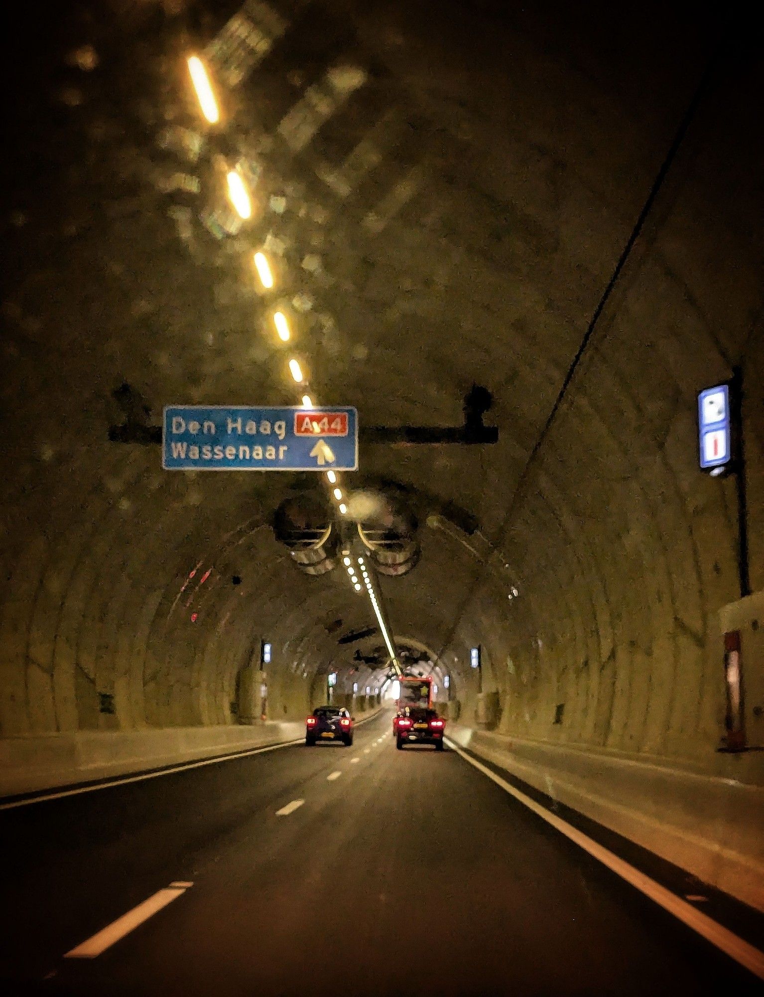 Bijna aan het andere eind van de Corbulo tunnel. Blauw bord richting Den Haag-Wassenaar. Regen op autoruit versterkt de lichten aan het plafond van de tunnel. Twee auto's in de verte.