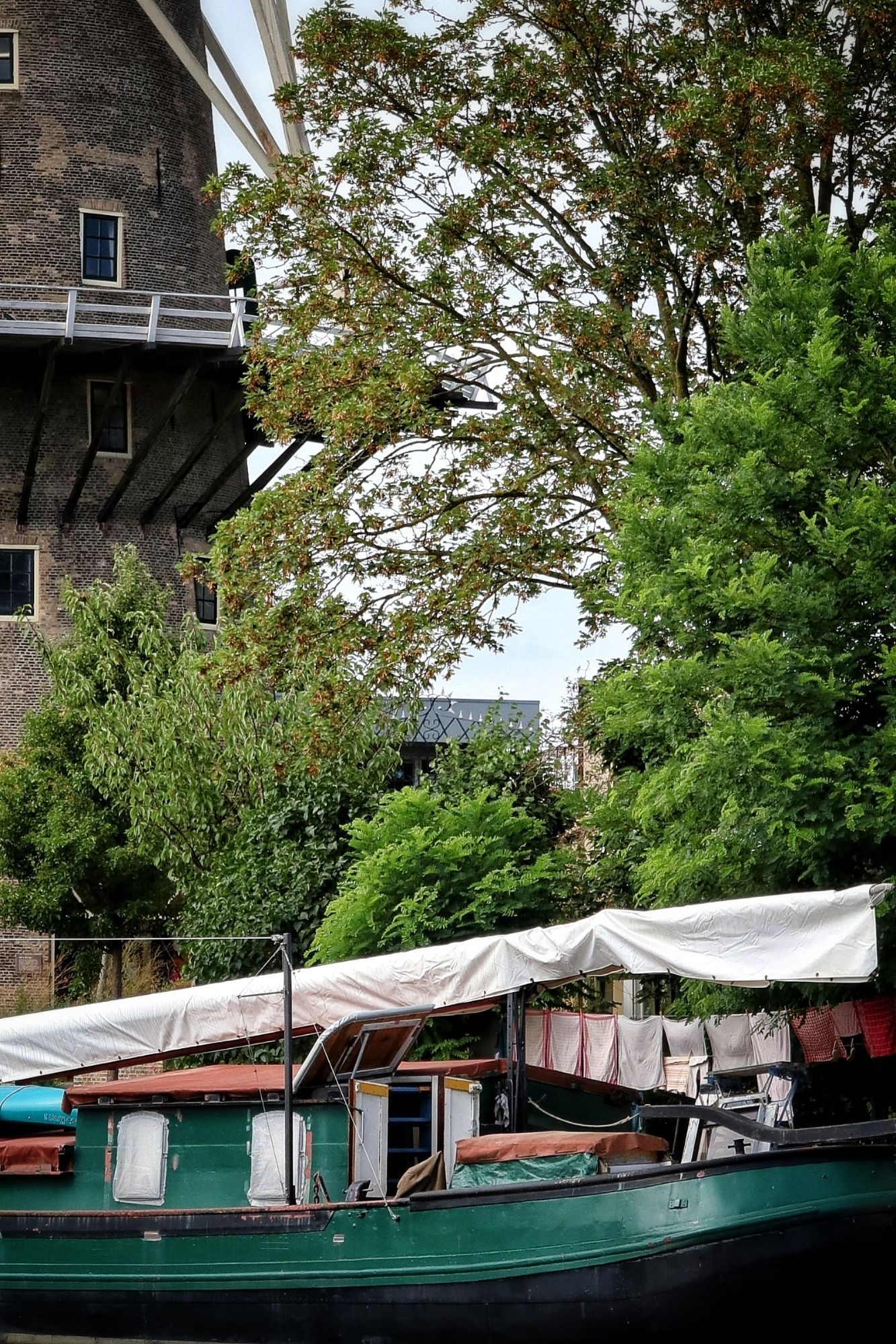 Was hangt te drogen aan een was lijn op een donkergroen geschilderde boot. Deel van bruin stenen molen zichtbaar op links. Bomen in vol blad achter de boot.