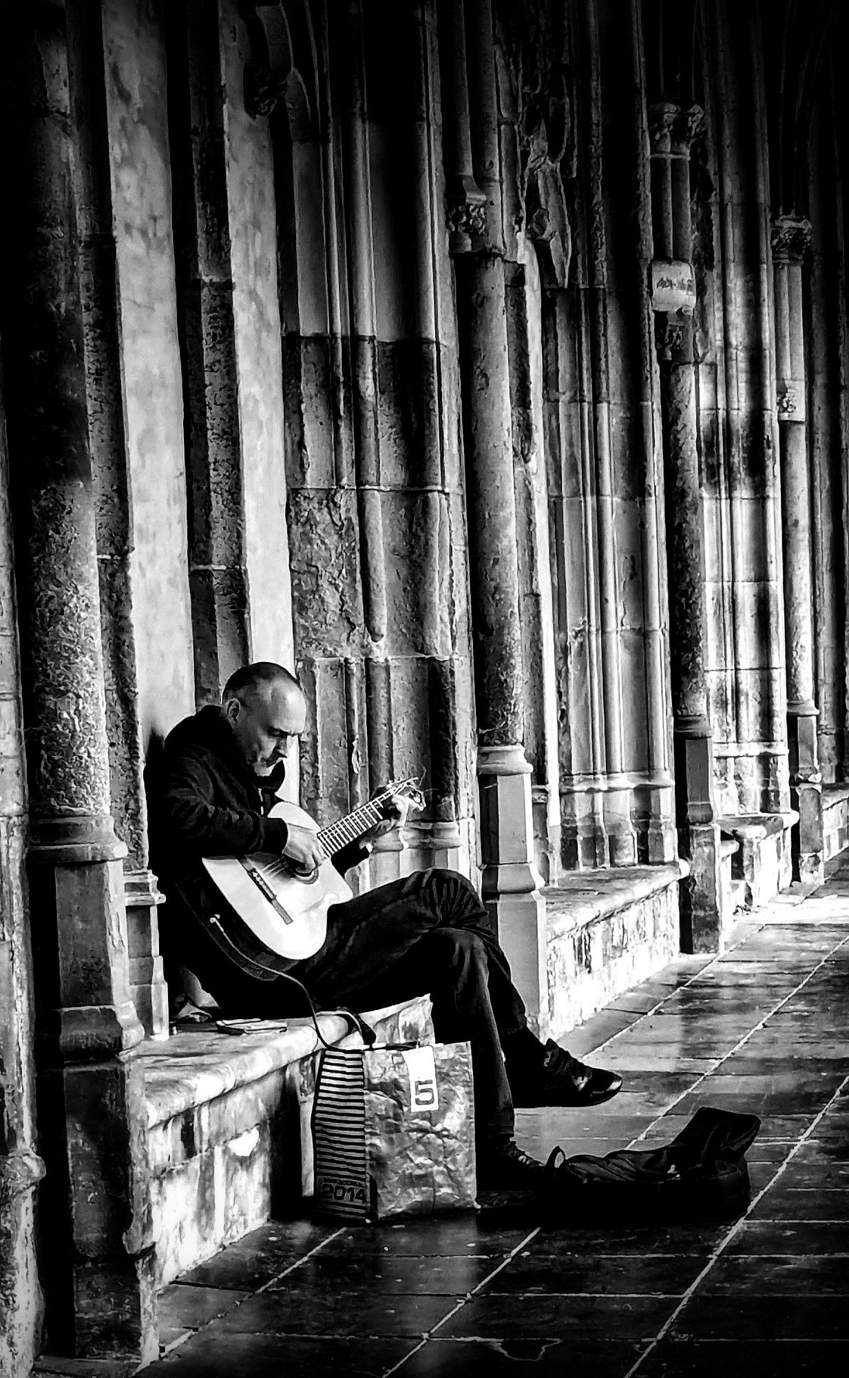 B&W. Man sitting down, softly strumming his guitar in an old cloister.