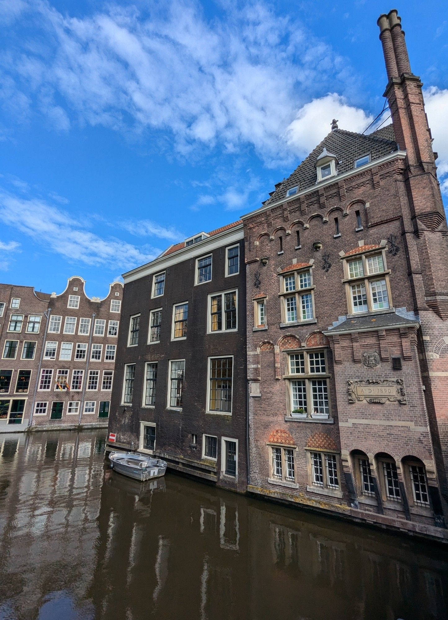 Grachtenpanden aan een gracht die naar rechts draait. Bootje voor één van de panden. Weerspiegeling in het water. Blauwe lucht met witte wolkjes.