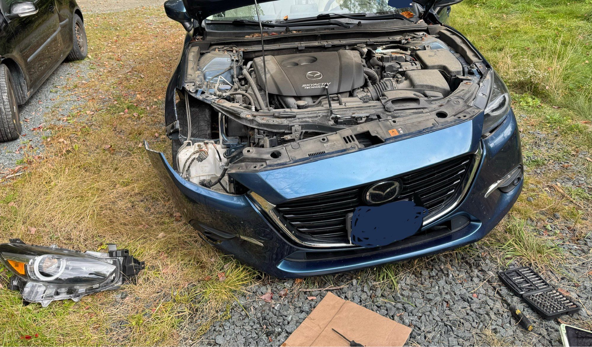 A blue Mazda 3 car with its hood open, the front bumper partially dismantled, and the passenger side headlight module removed