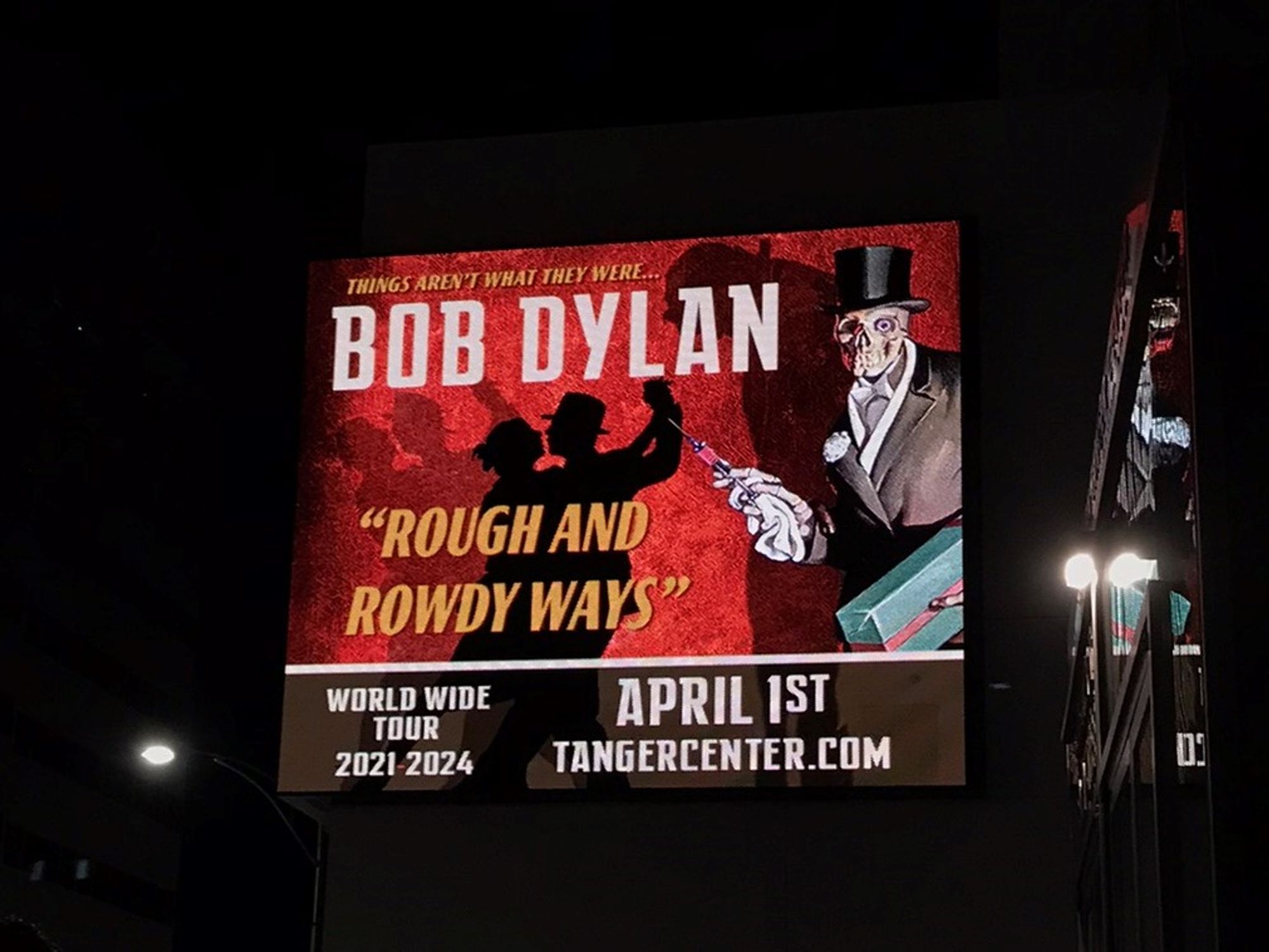 A lighted sign in the nightime dark.  Red background with a couple dancing and a skeleton wearing a top hat and holding a syringe and a wrapped gift. "Things Aren't What They Were...Bob Dylan World Wide Tour 2021-2024."