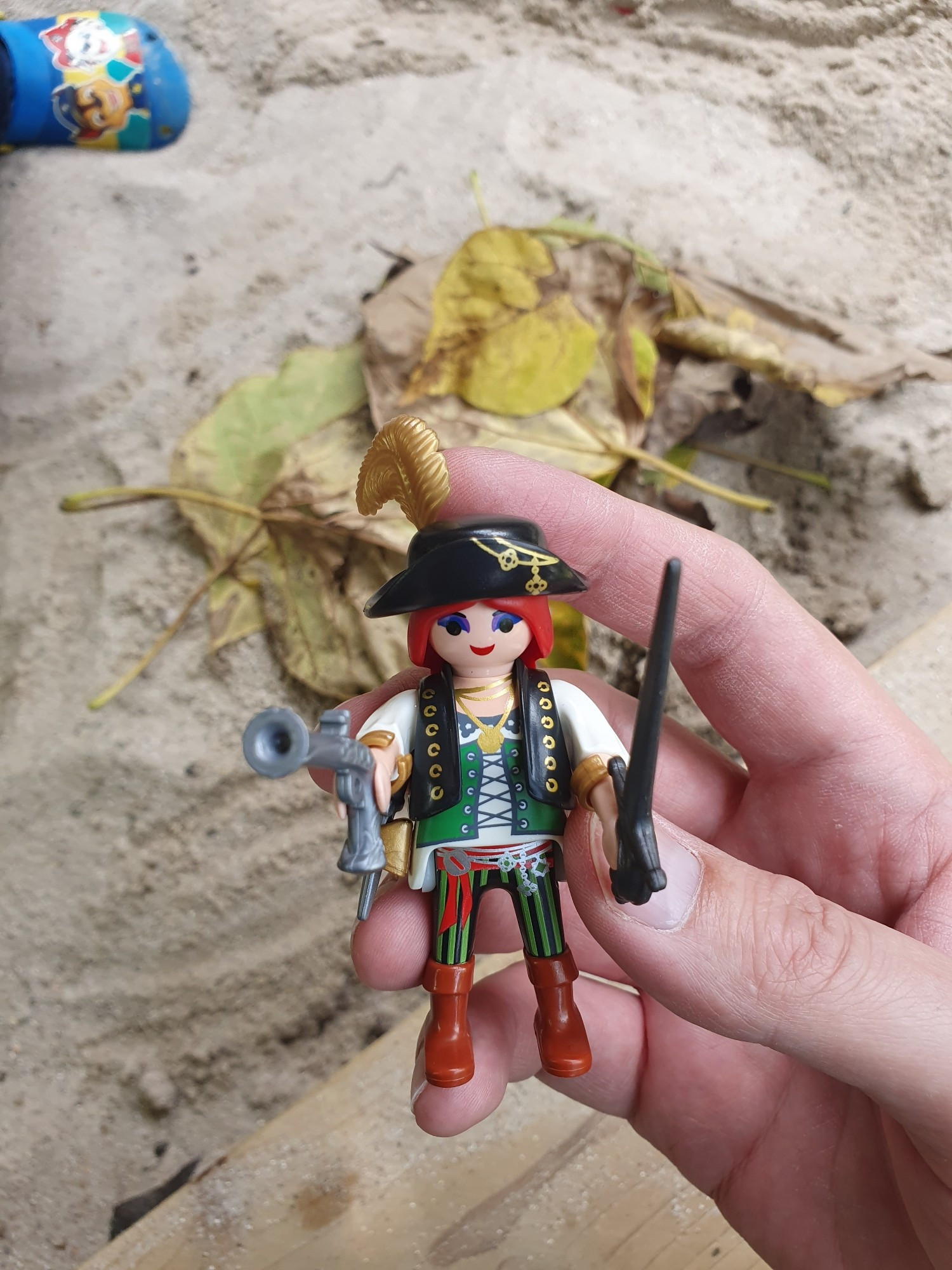 Ich halte in meiner rechten Hand eine rothaarige Playmobil Piratin.
Die Piratin trägt einen schwarzen Hut mit goldenen Schmuck. Dazu ein weisses Hemd mit einer grüne Weste und eine schwarz und grün gestreifte Hose. In den Händen hält sie ein Schwert und eine Pistole.