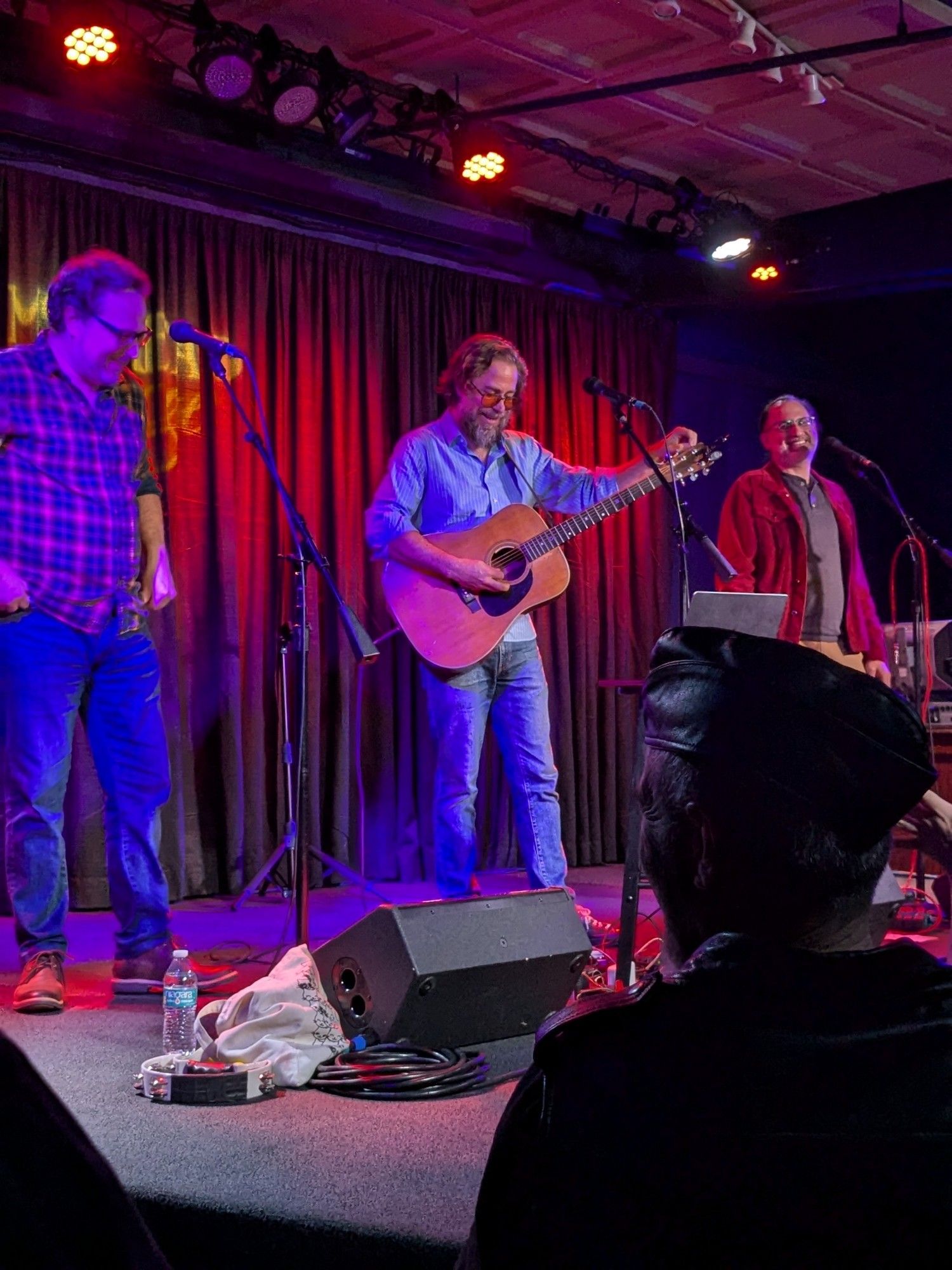 Jonathan Coulton on a small stage, tuning his guitar, flanked by Paul and Storm.