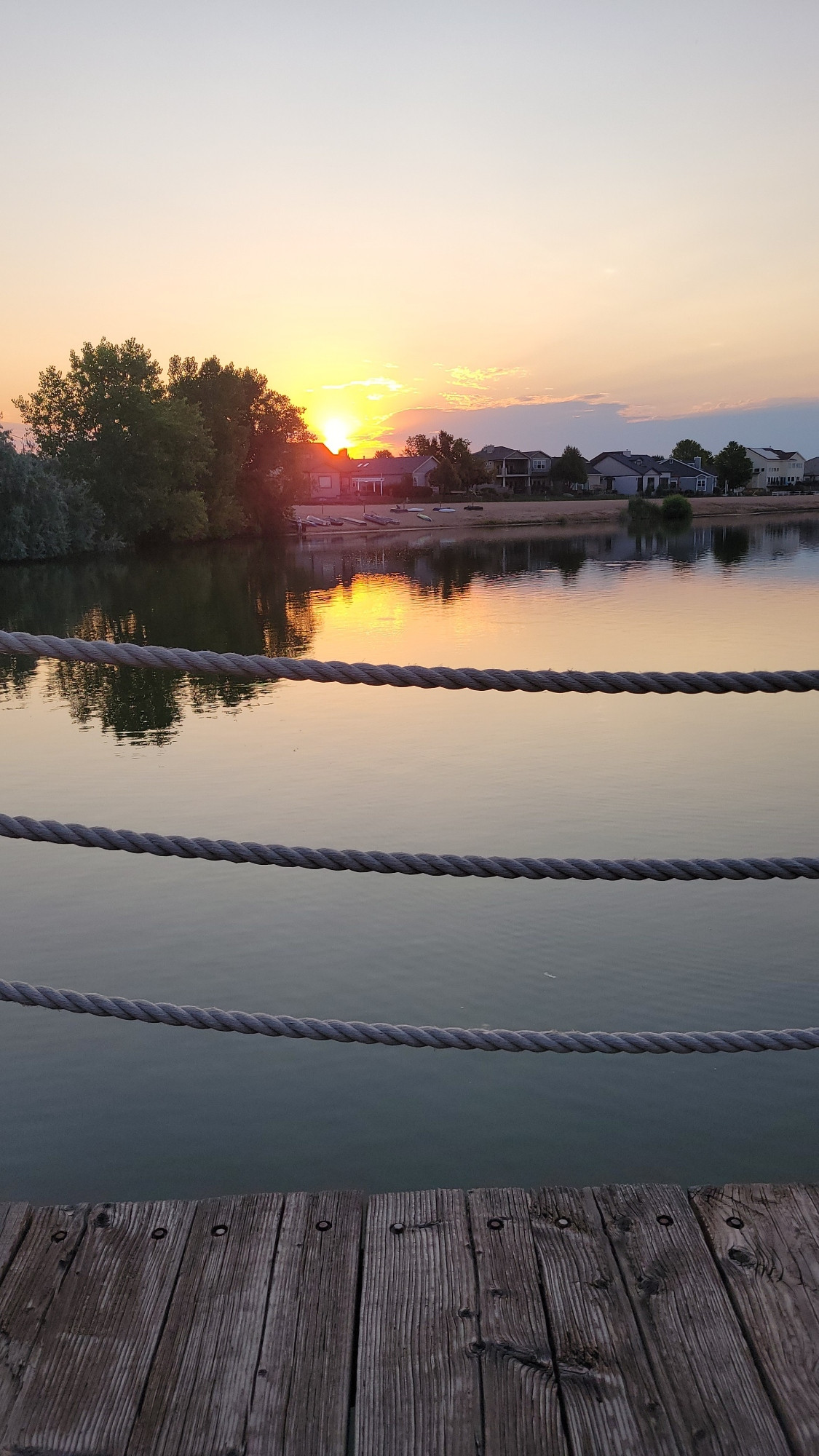 Boardwalk with rope fence in foreground, calm glassy lake reflecting the trees and sunrise, low clouds on the horizon