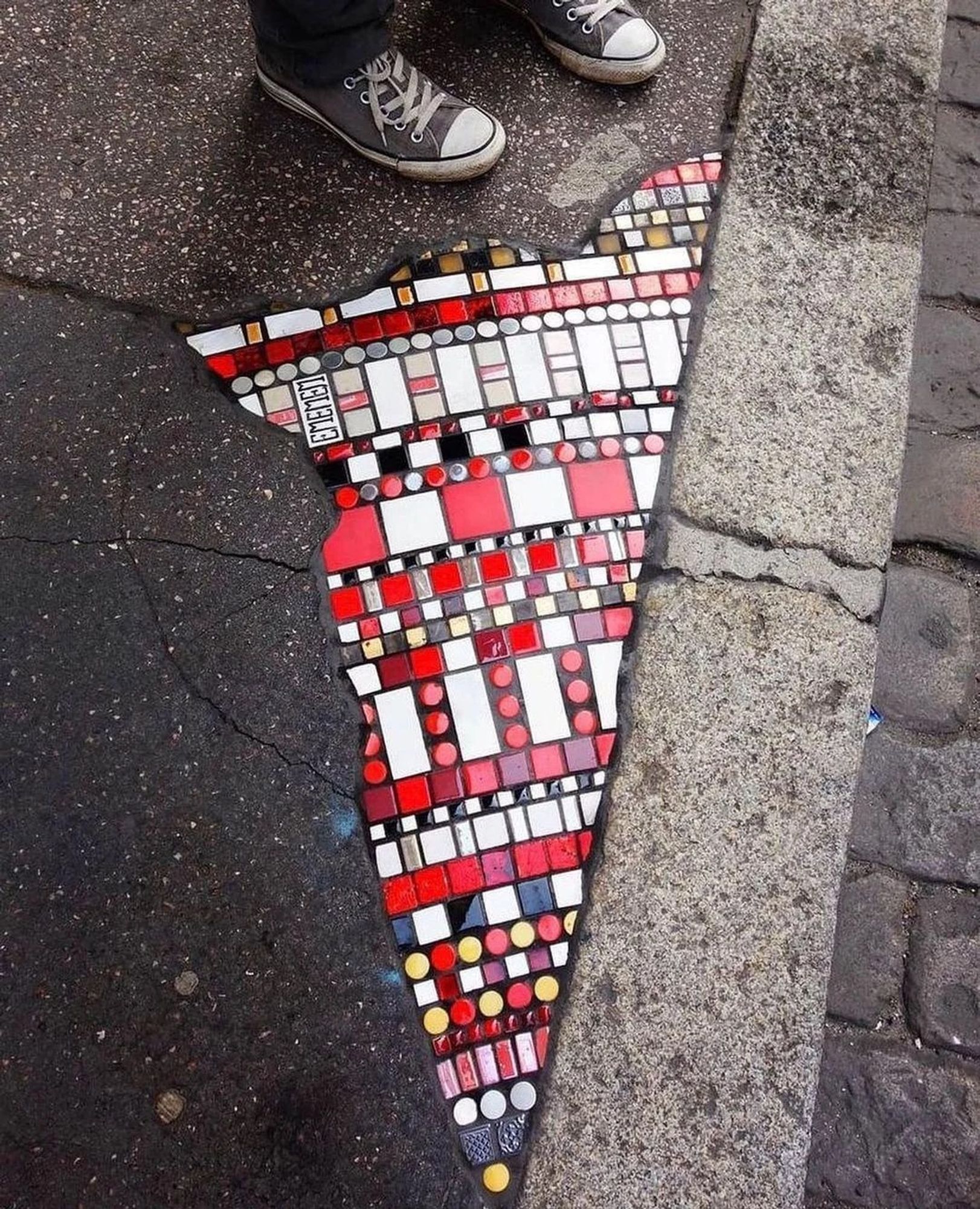 mosaico com várias peças redondas, quadradas e retangulares em vermelho, amarelo, preto e branco, formando um padrão que lembra muito a arte indígena, em uma parte de uma calçada, num formato quase triangular.