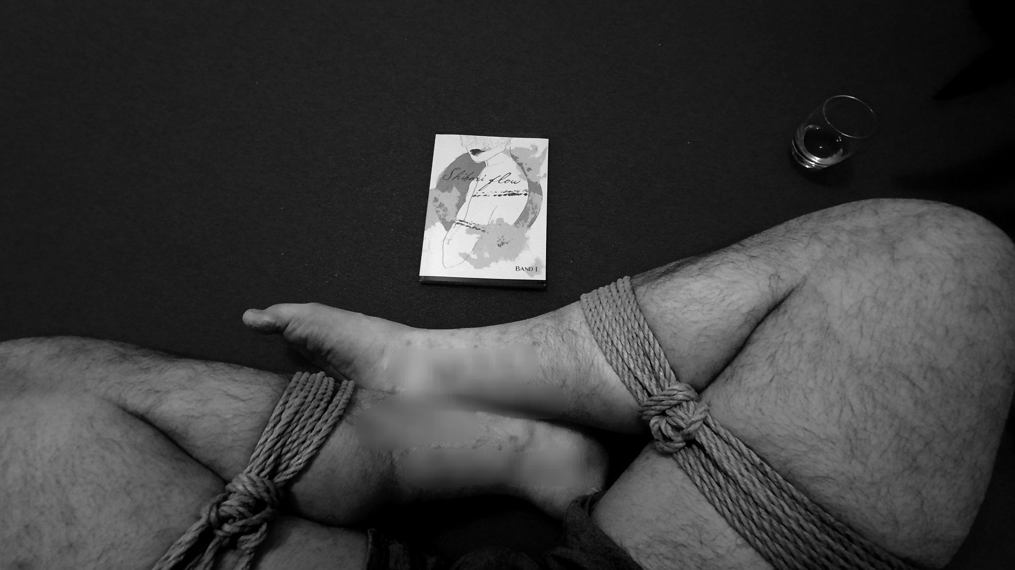 Selbstfesselung im somewhat-frogtie, Whisky, Shibari Flow Magazin als Lektüre. Bild in Schwarz-Weiß