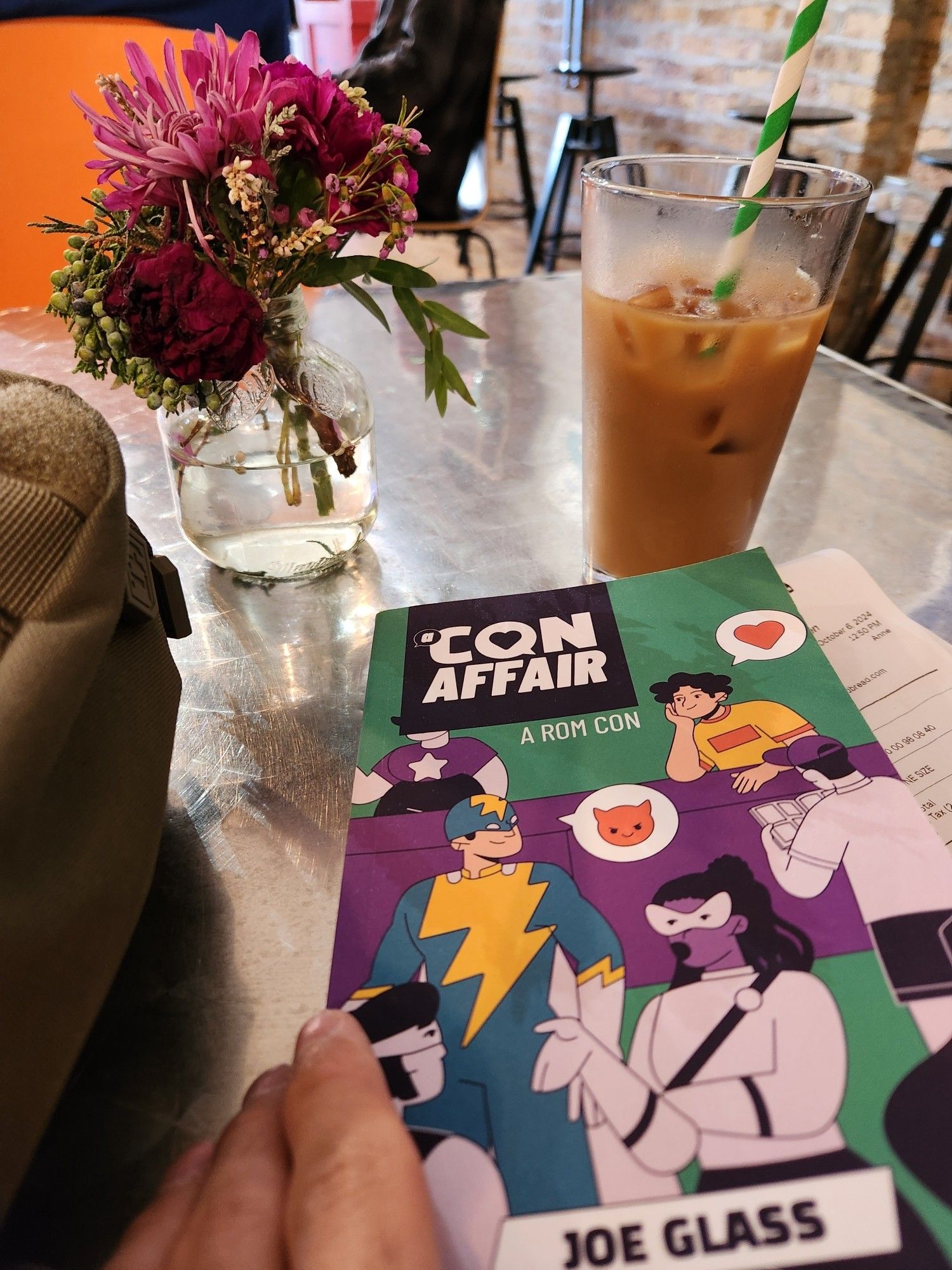 A photo of a small vase of flowers next to an iced coffee with a booked called A Con Affair laid out on a Cafe table