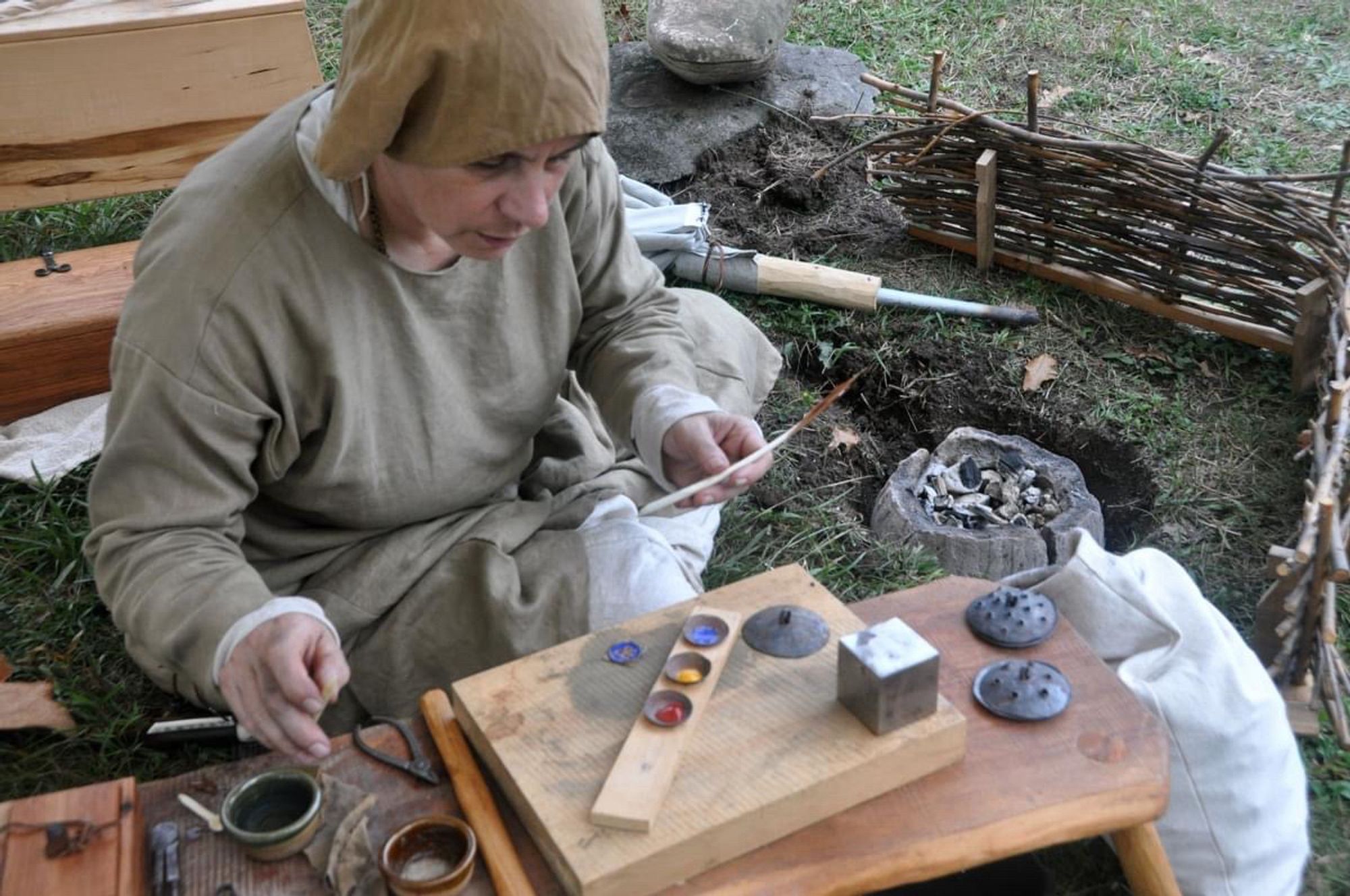 A woman in Anglo-Saxon clothing makes enamelled pieces next to a small charcoal fire.