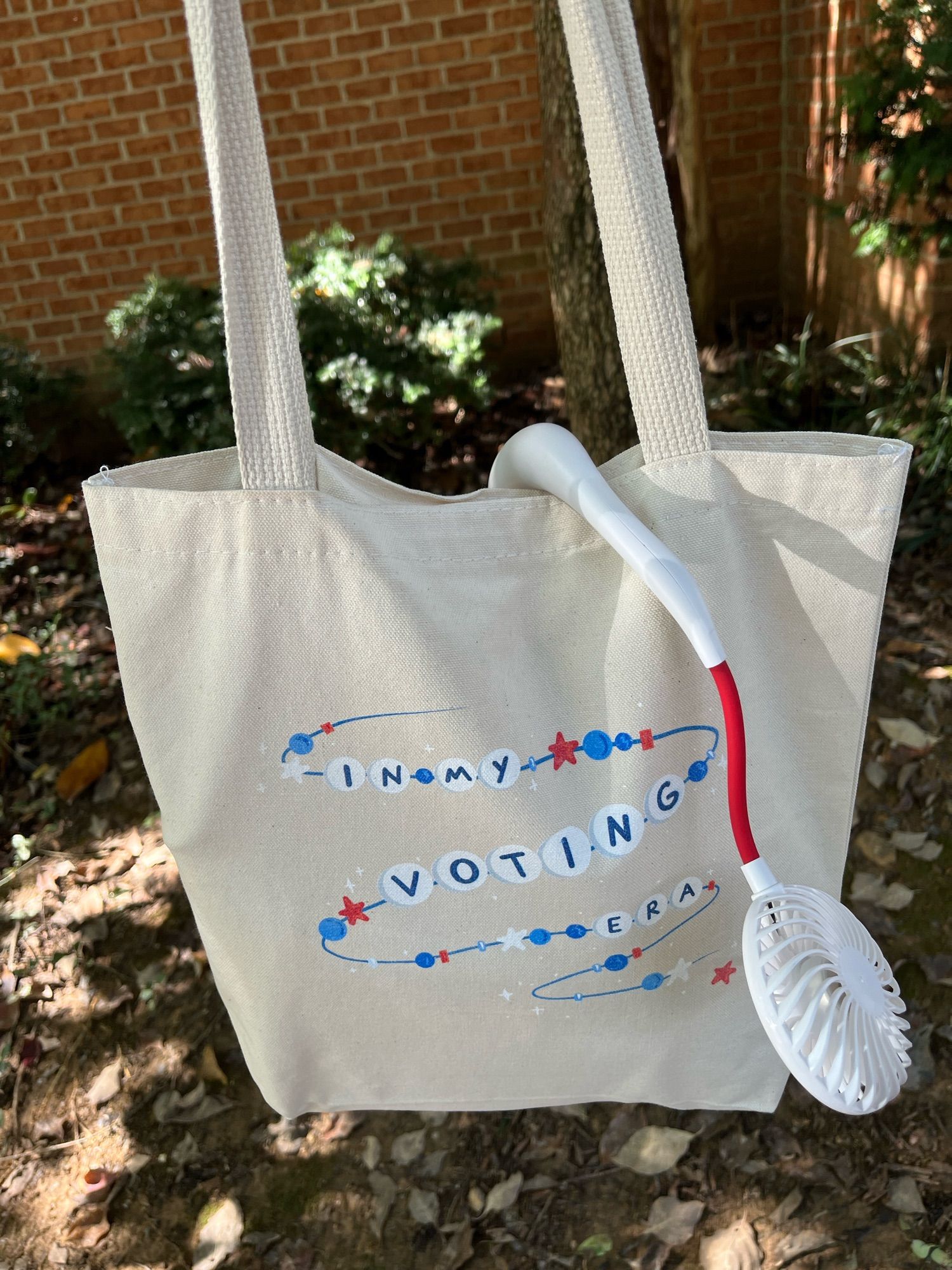 A beige canvass tote bag that says IN MY VOTING ERA on a friendship bracelet, in blue and white and red. A plastic USB neck fan hangs across it.