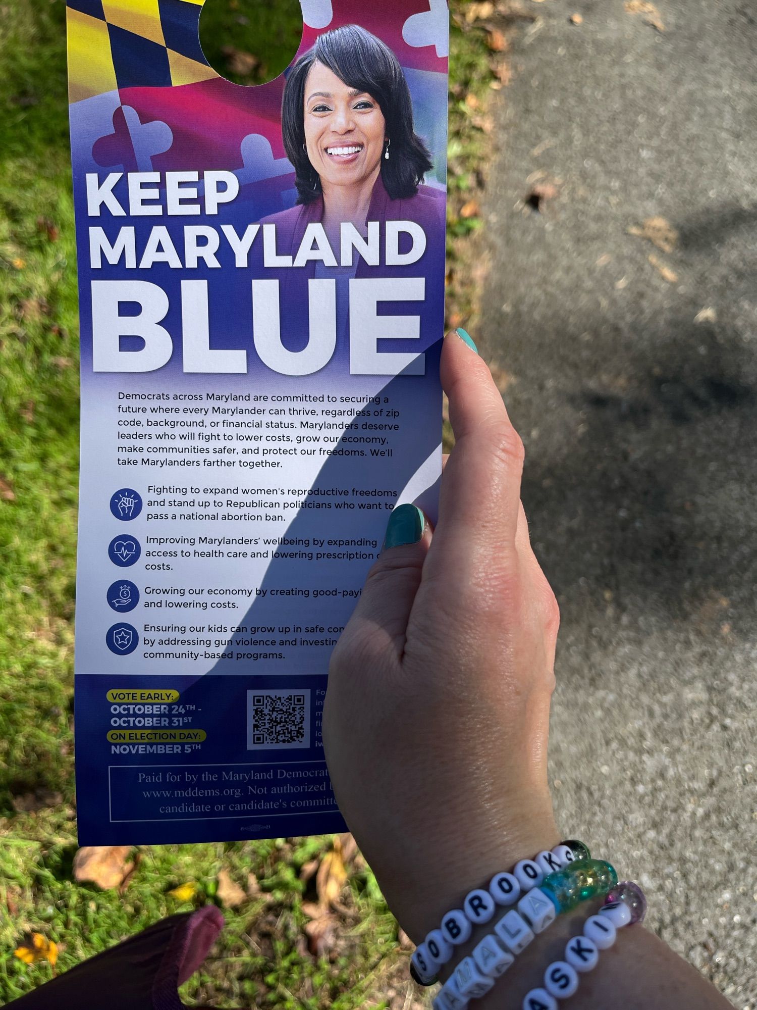 A hand with friendship bracelets that say Alsobrooks, Kamala, and Jamie Raskin holds a piece of Keep Maryland Blue campaign literature for Angela Alsobrooks.