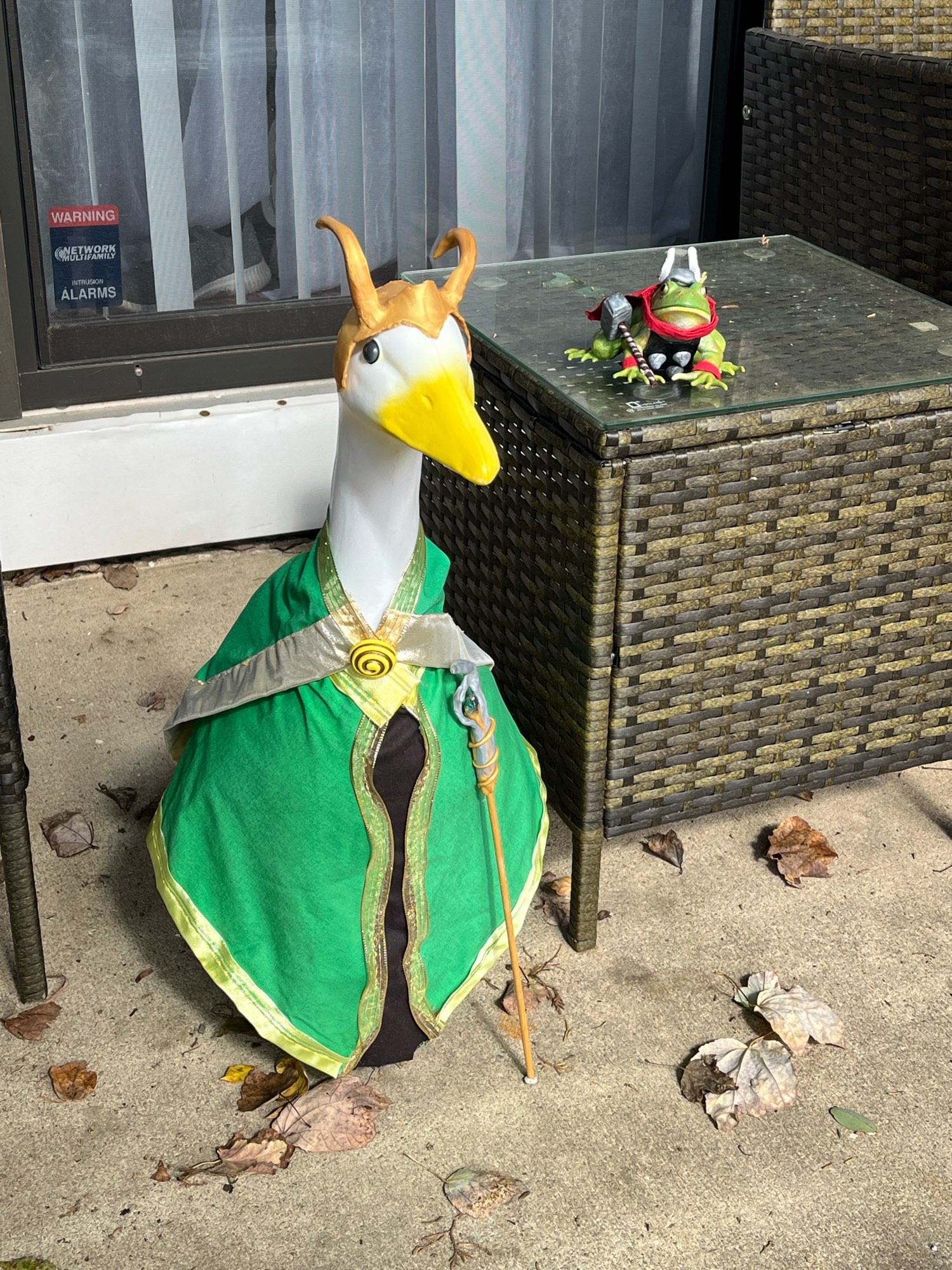 A white plastic porch goose dressed as Loki with a green, black and gold outfit and scepter and horns sits on a patio next to a brown wicker table. On the table is a green plastic frog dressed as Thor.