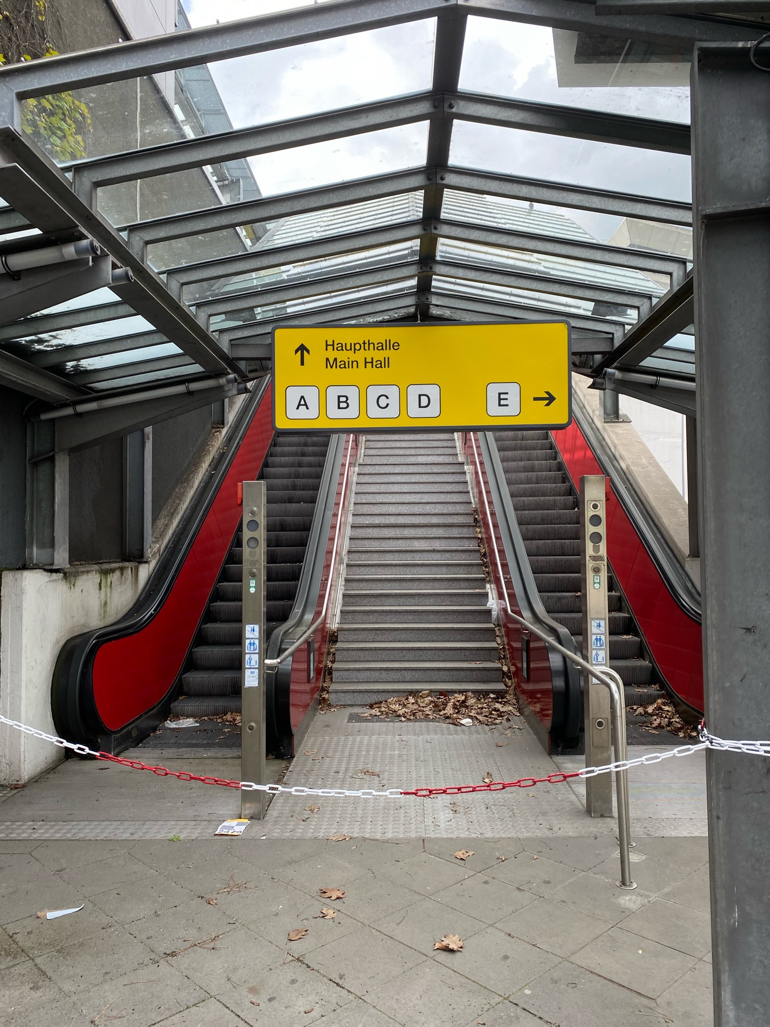 Stillgelegte Rolltreppe, mit rot-weißer Kette versperrt. Eine Treppe zwischen den Rolltreppen, in einer Halle mit Glasdach