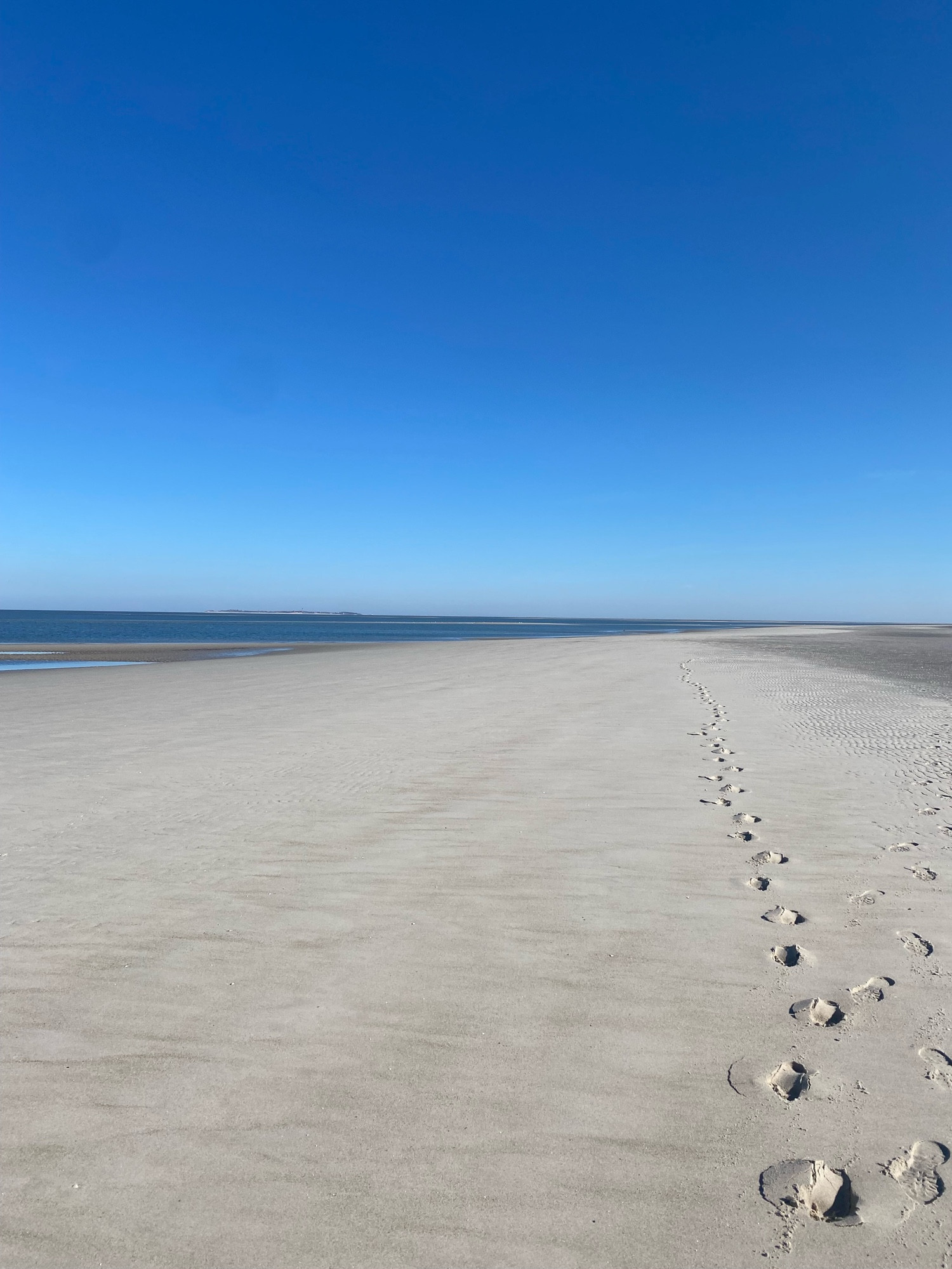 Ganz viel Sandstrand, etwas Meer und blauer Himmel, Fußspuren im Sand.