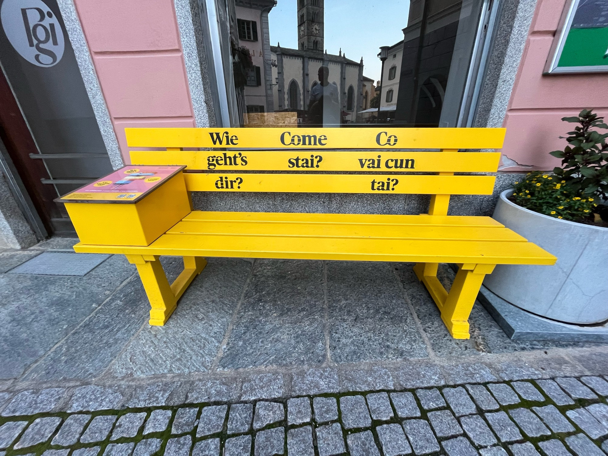 Gelbe Bank mit Schriftzug „wie geht’s  dir? Come stai? Co vai cun tai?
Dahinter ein Schaufenster mit Spiegelung einer Kirche. 