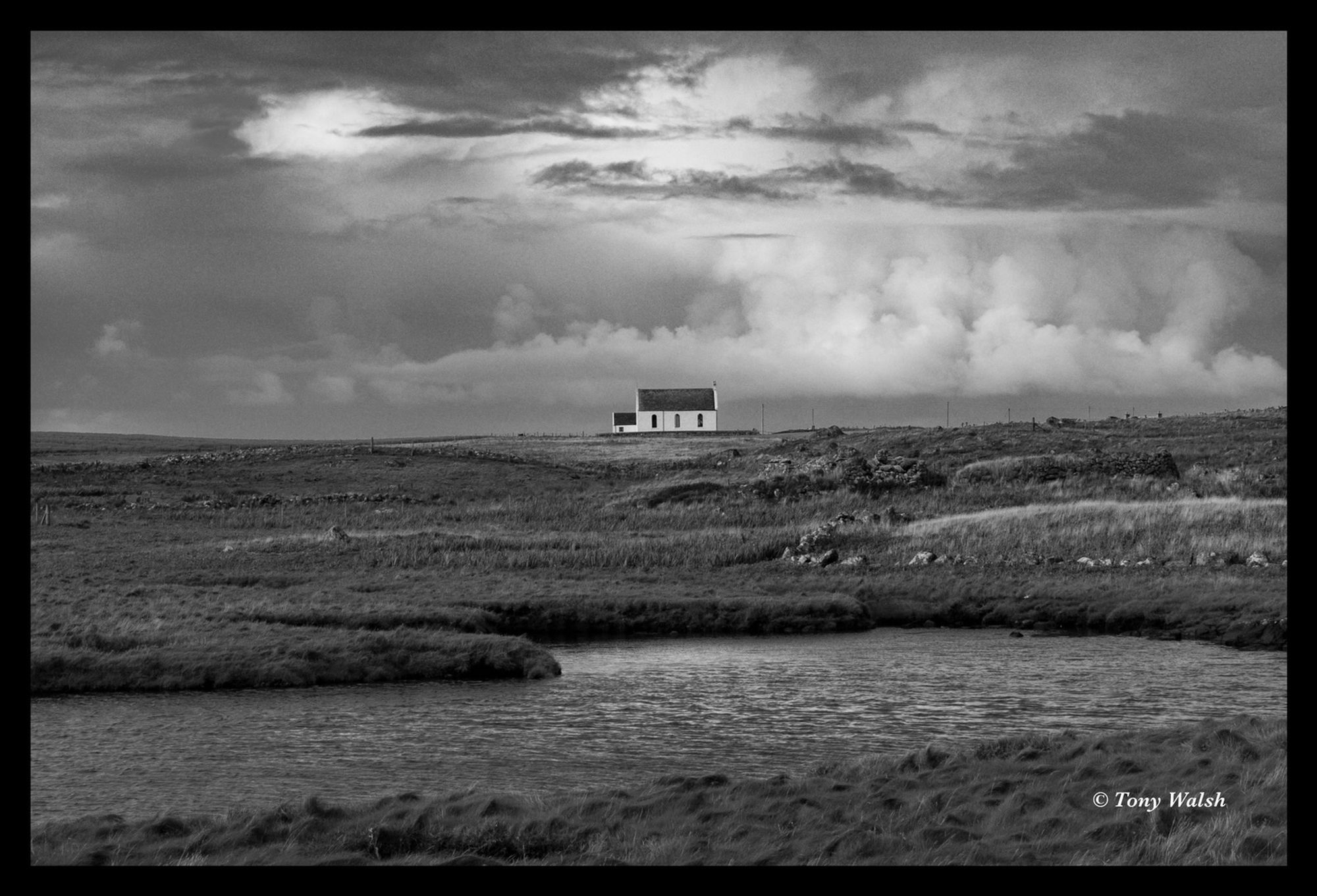 Black and white image of a small white church in the distance with clouds behind it and a small loch in the foreground