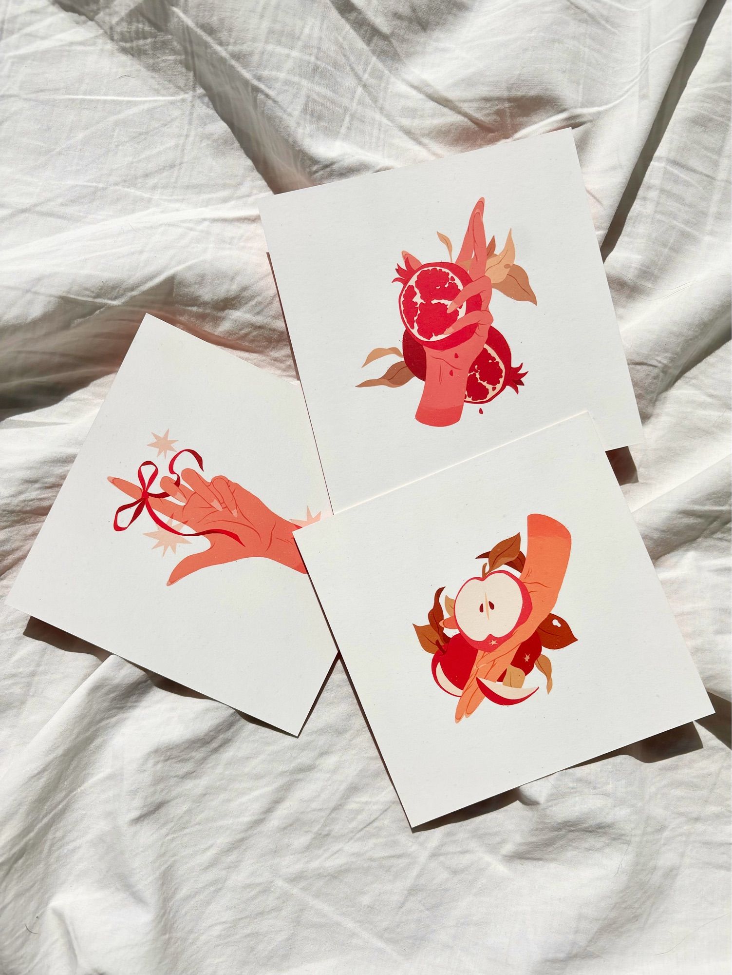 Three mini prints of a medium pink hand holding a pomegranate, a light orange hand holding an apple, and medium peach hand with a red ribbon its finger. All three on a white sheet background