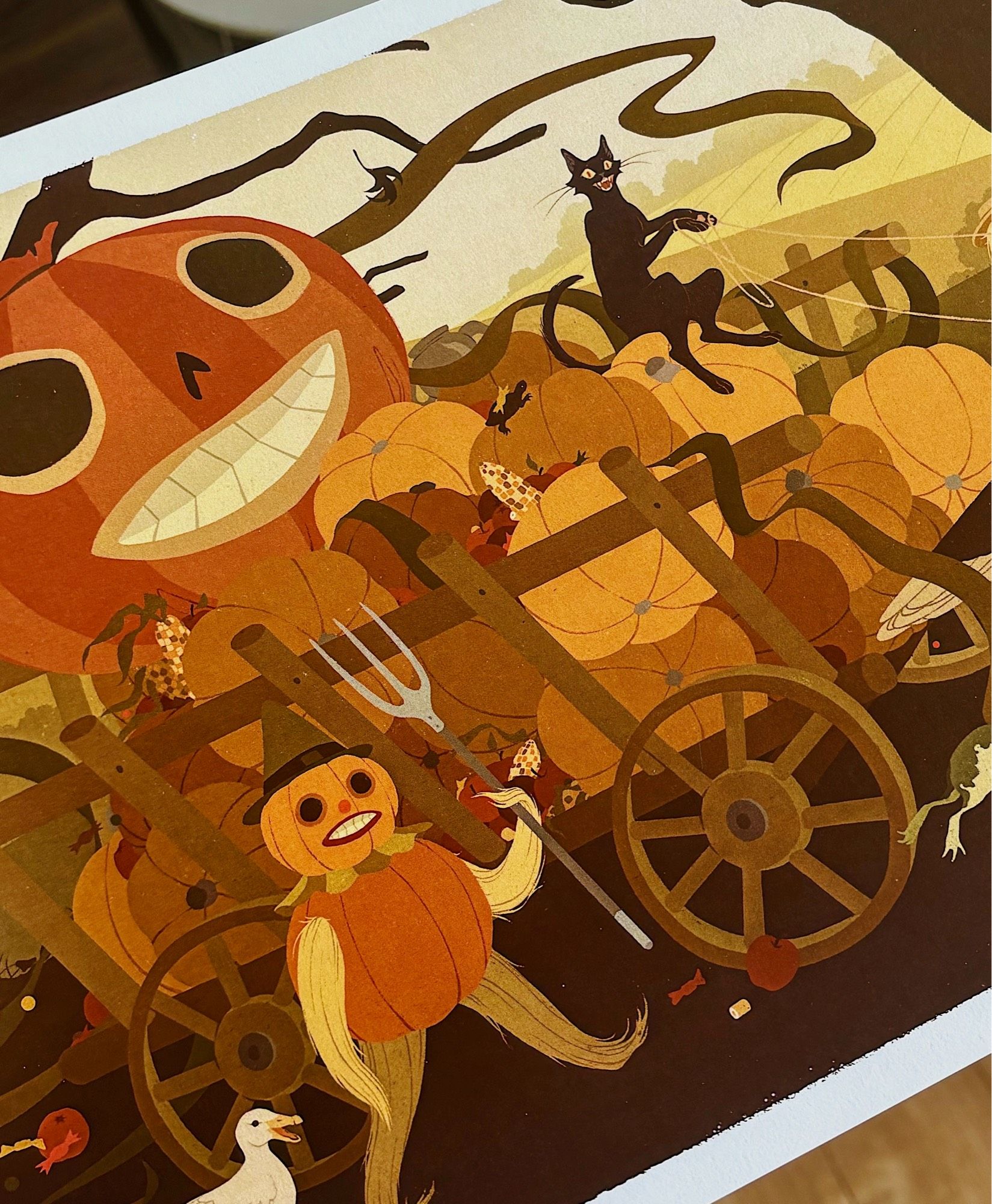 Crop of a print of fan art for Over the Garden Wall. Shows a cart with pumpkins being ridden by a black cat and a pumpkin man with a pitchfork walking next to the cart. The background is of a bucolic scene with rolling fields