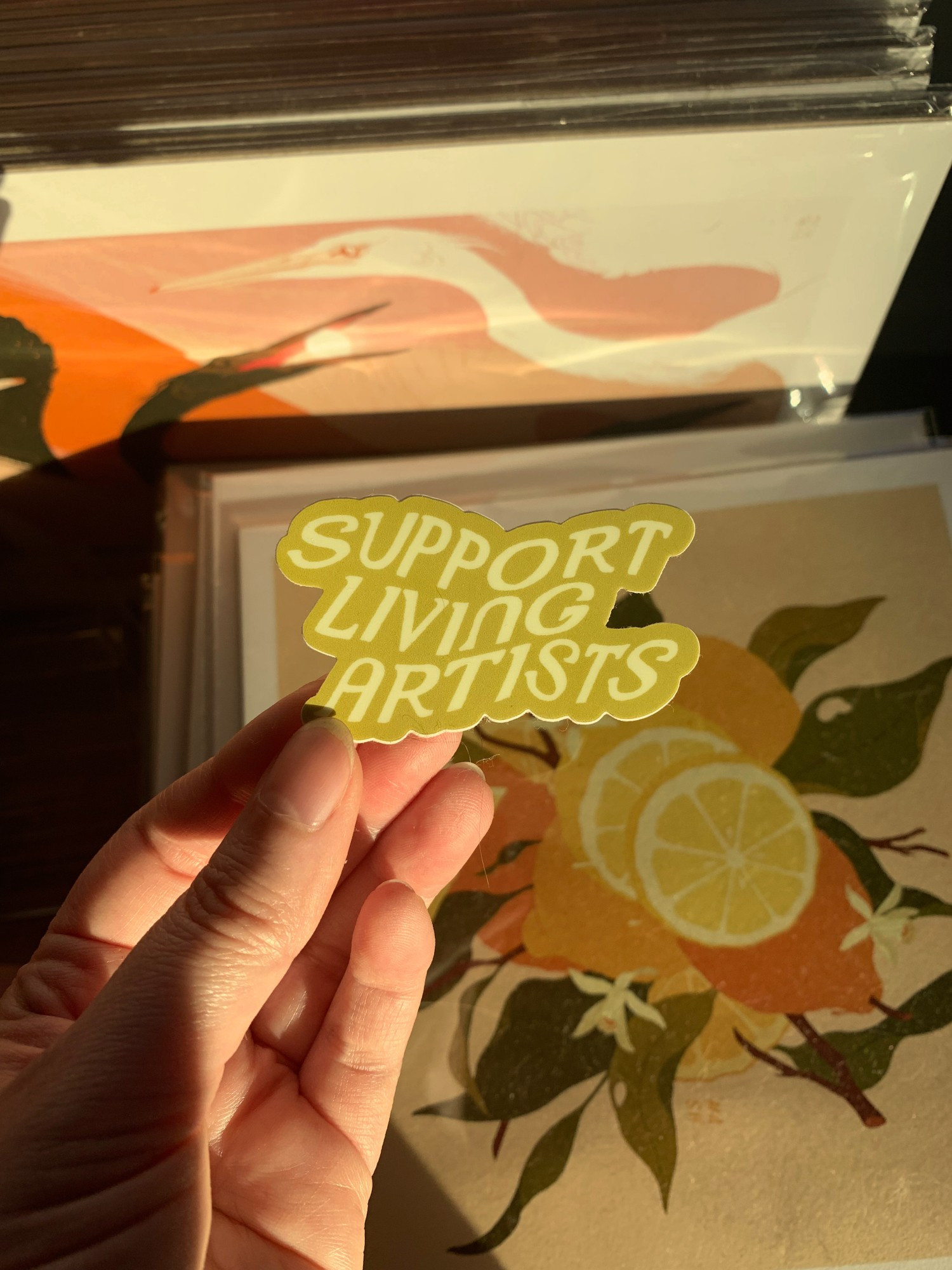 Samantha Mash Illustration sticker being held by a pale hand. The sticker is a chartreuse green and cream color with the text being lighter, it says "Support Living Artists"