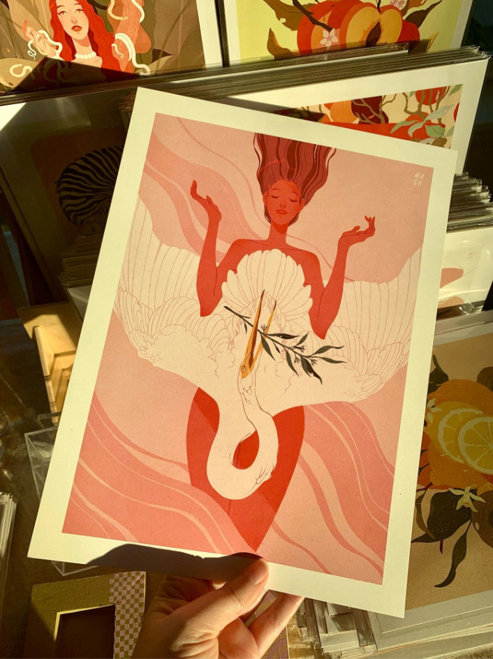 Samantha Mash Illustration print being held by a pale hand. Print is of a woman with deep pink skin and slightly darker hair holding her her arms up with with a light pink background. A white crane rests over her chest and lower body holding an eucalyptus branch