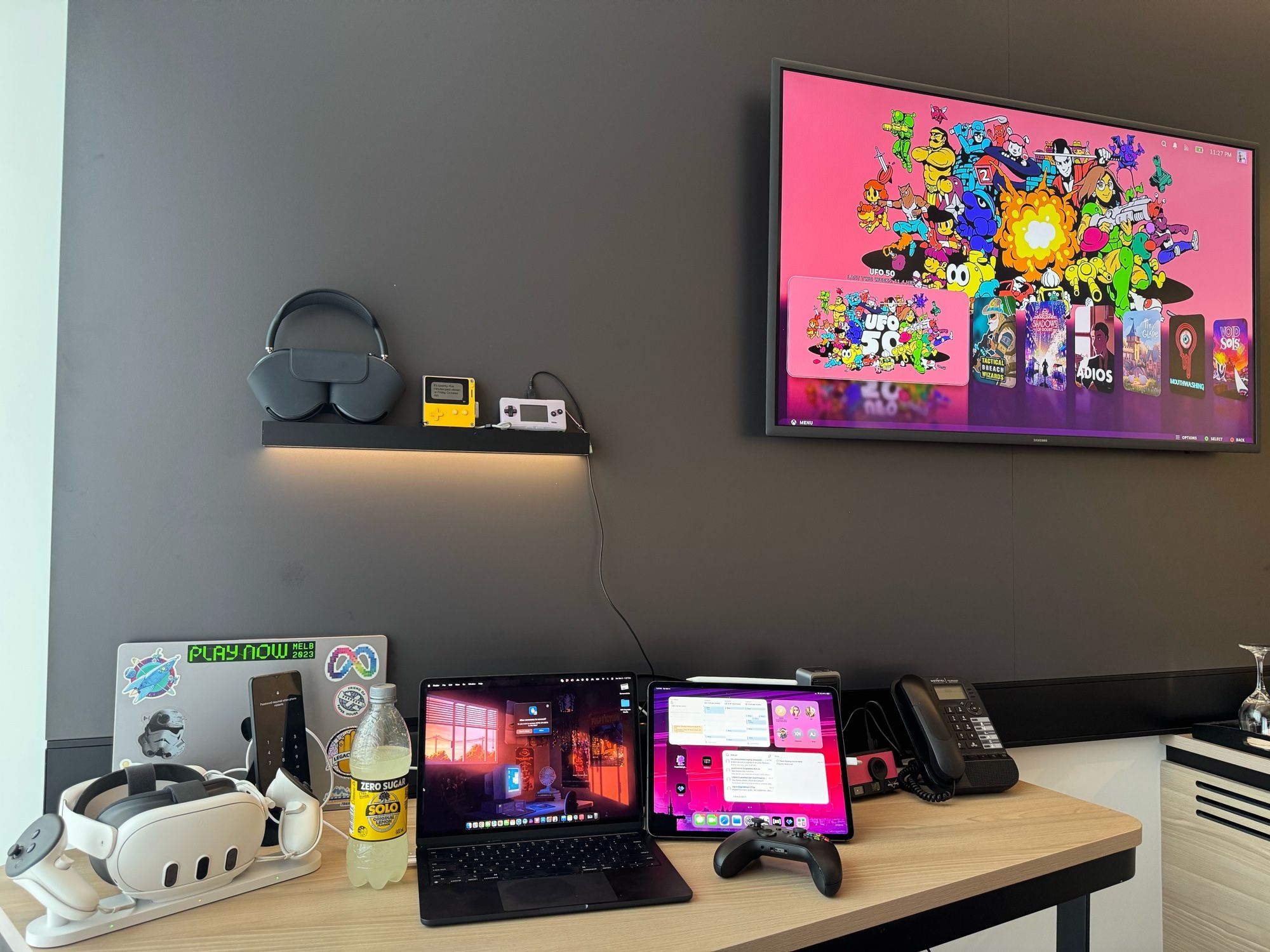 My hotel desk. Right to left: A charging Quest 3 and cell phone in front of a closed MacBook (my work one), an ops MacBook Air and an iPad Pro. A charging battery and a charging CampSnap camera.

Above: AirPod Max headphones, a Play date showing its clock face, a charging gameboy micro.

The TV is showing my Steamdeck (out of sight behind the other things), with UFO 50 highlighted