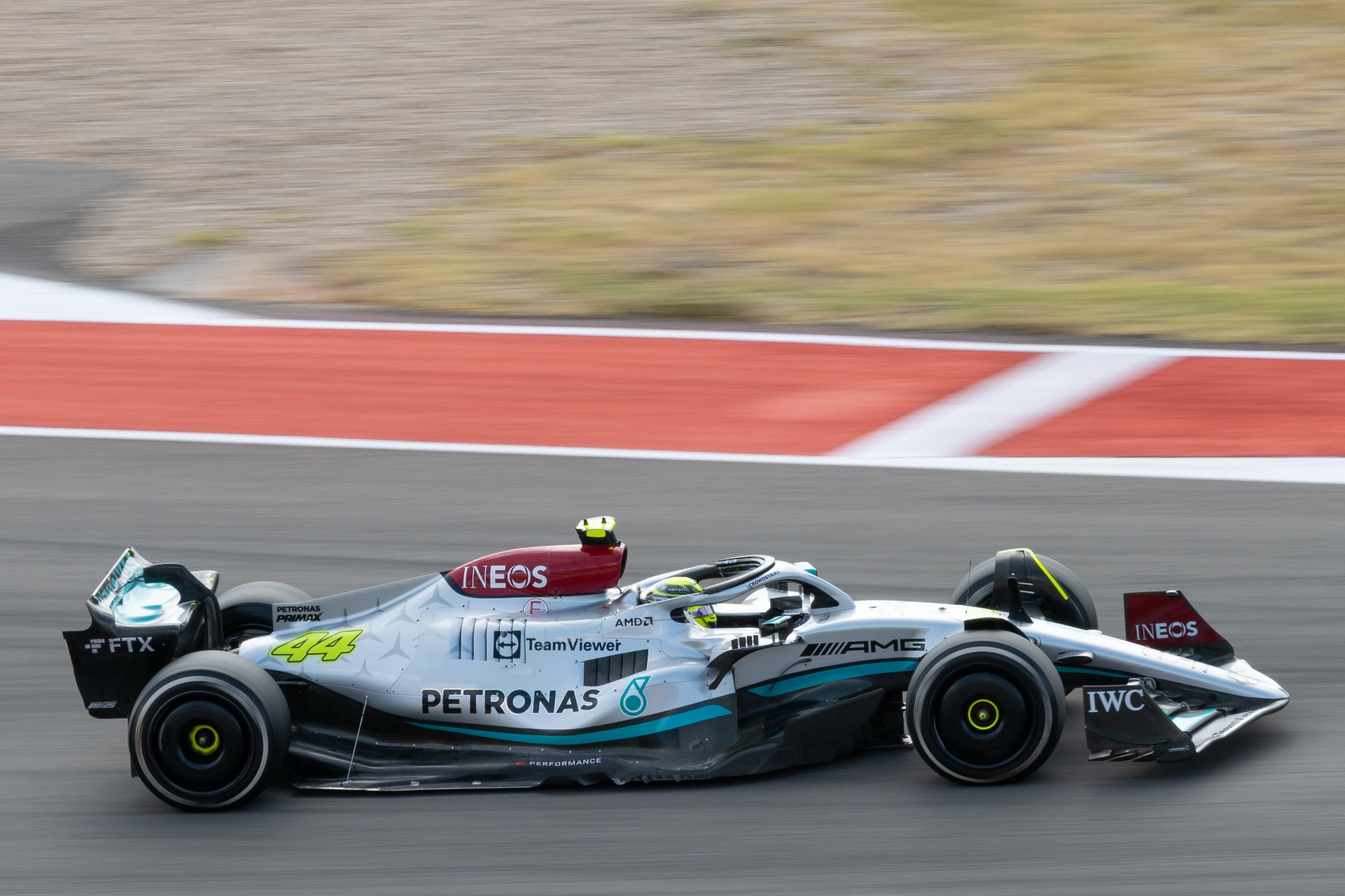 Mercedes W13 Formula 1 car in Lewis Hamilton's livery: Mostly silver, with a wave of black at the bottom, separated by a blue streak. The air intake and inside of the front wing are a deep red; the number 44 on the engine cover, the camera pod above the air intake, and Lewis' helmet are yellow. The car is in focus, but the background is blurred: pavement in front, red and white curbing above, and brown/yellow grass in the background. 