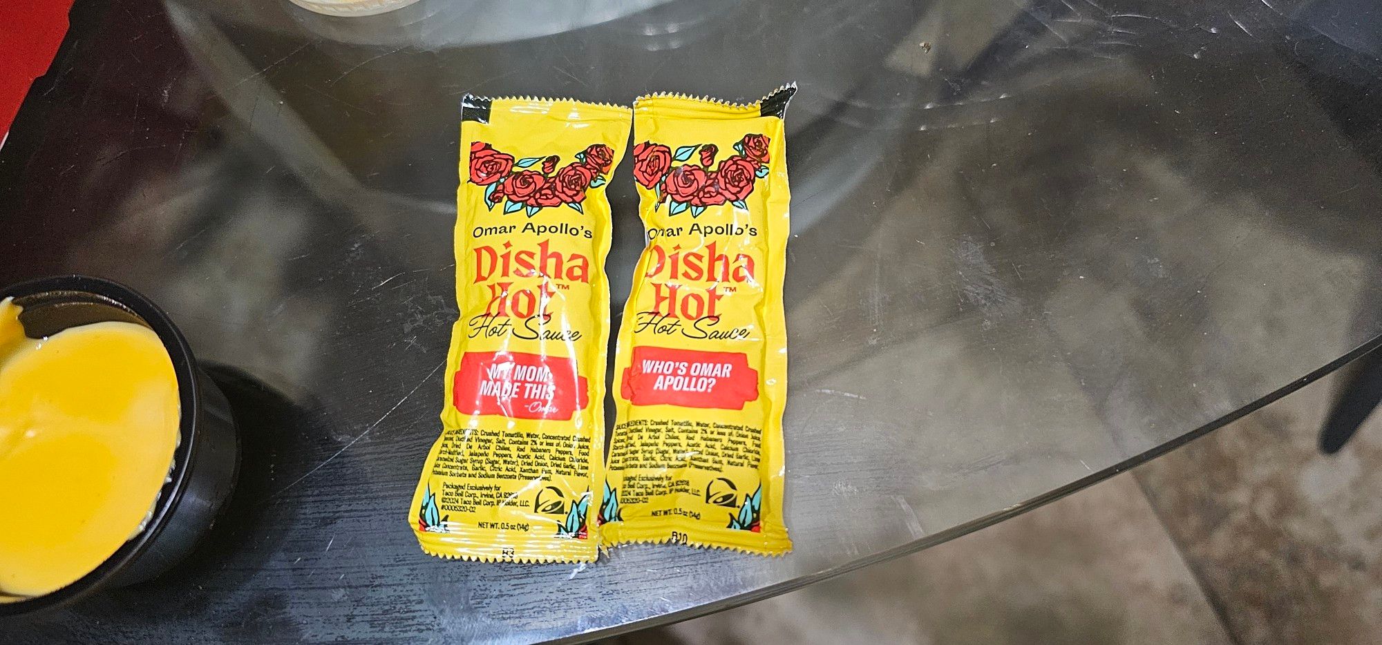 Two bright yellow packets of Omar Apollo's disha hot sauce, one packet says "my mom made this" and one says "who's Omar apollo". I got cup of nacho cheese in the picture for no reasons too