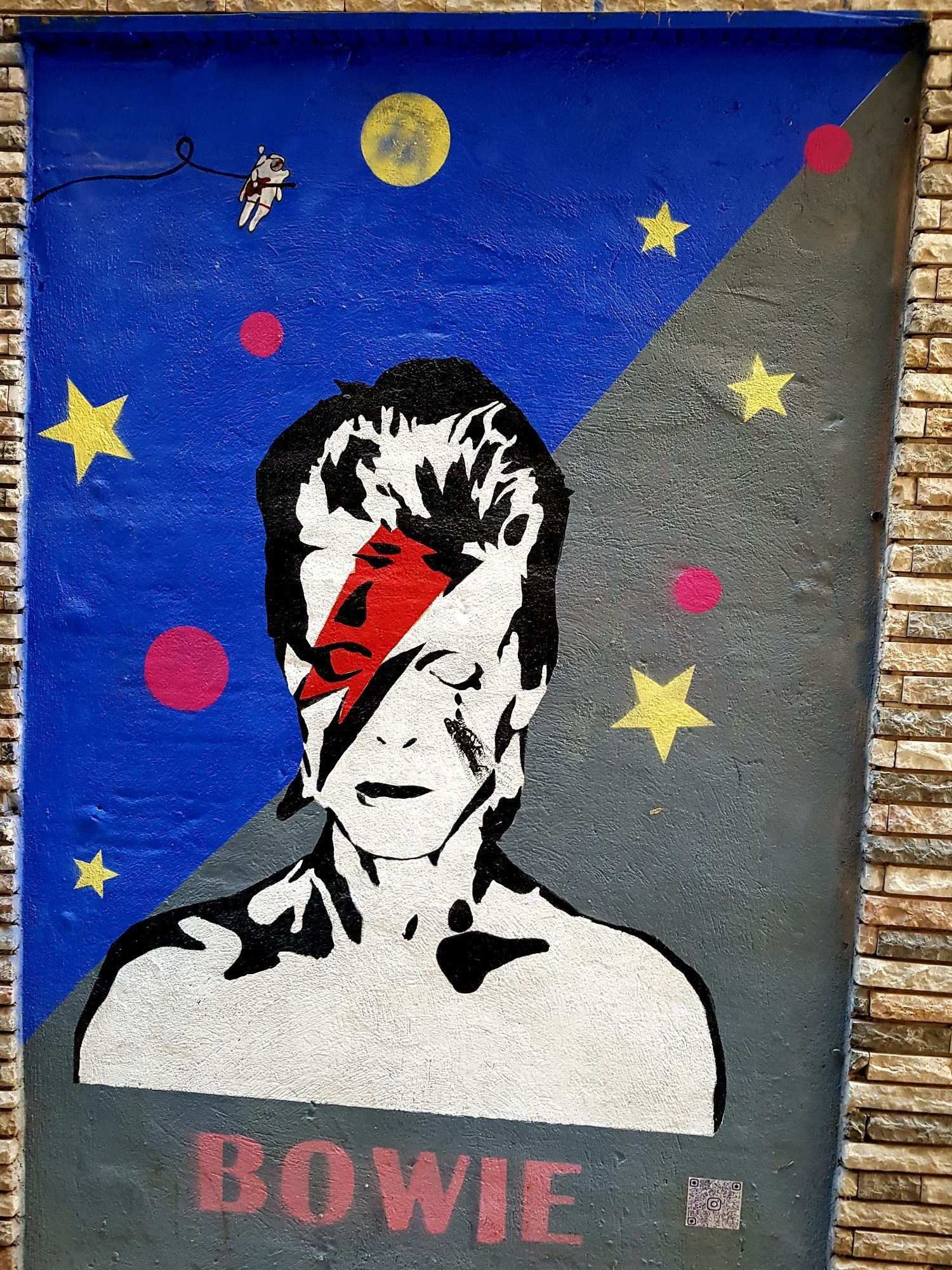 Graffiti. Abgebildet ist David Bowies Torso, schwarz und weiß. Auf der Stirn ein roter Blitz. Links und rechts von ihm sind Sterne und Kreise.