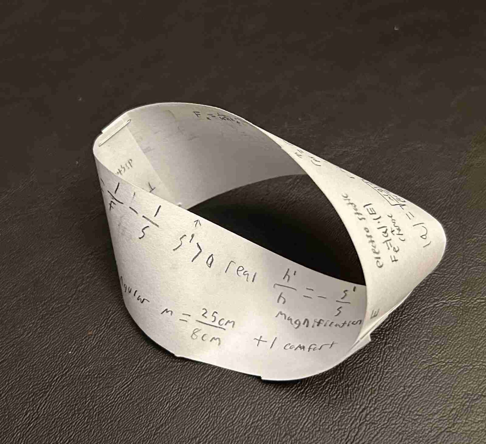 A three by five inch piece of white paper has been cut in half lengthwise and stapled back together with a twist to form a Möbius strip. Physics equations and reference information are written on all surfaces, but it only has one side.