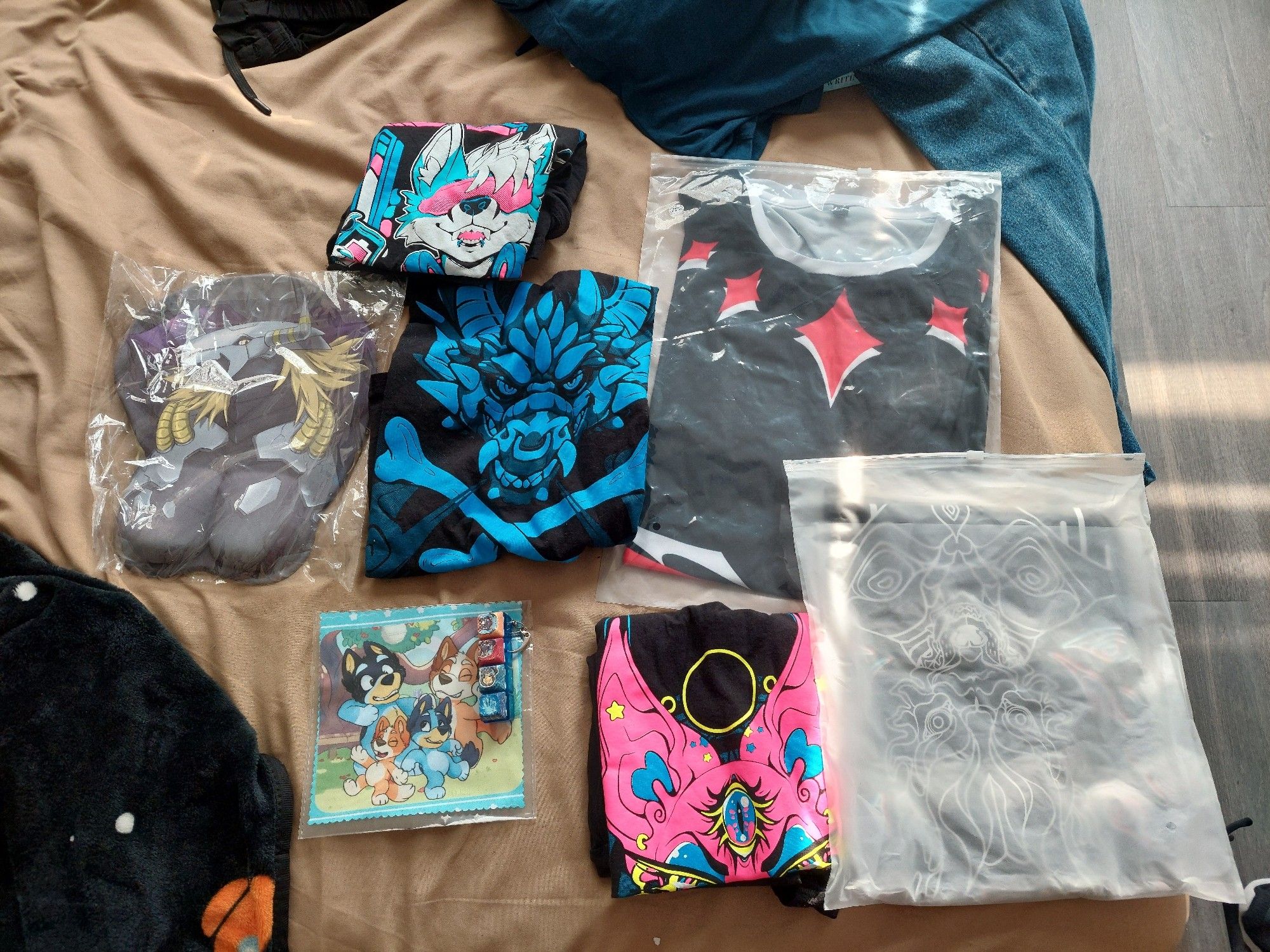 Strwen across the bed are items purchased from StratosFur's Dealer's Den. In total, there are eight (8) items being shown. Five clothing items and three accessories.

Three (3) Tank Top Shirts - Dead Bomb Art
One (1) Graphic Tee Shirt - SanityisFURtheSane
One (1) Double-Sided Shirt - SanityisFURtheSane
One (1) BlackWarGreymon Oppai Mouse Pad - SanityisFURtheSane
One (1) Bluey 'Dance Mode' Microfiber Cleaning Cloth - Moko's Makery
One (1) Luxray Clicky Key Fidget Toy - Moko's Makery