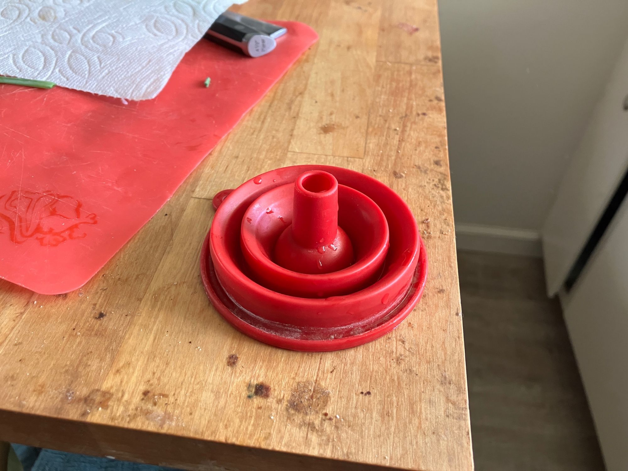 A red collapsible silicon funnel on a wooden board, it is mostly collapsed with just a little nub standing up reminiscent of a cold water shrunken penis.