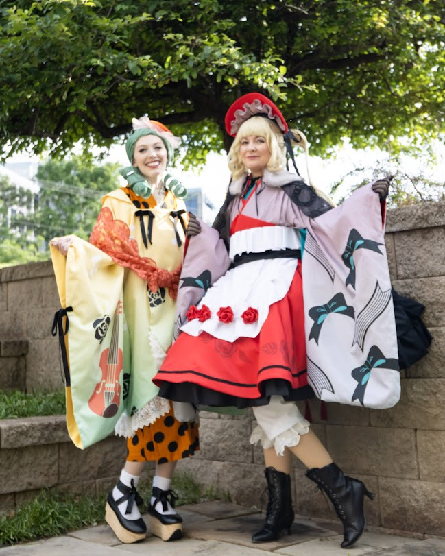 Dahlia wearing a yellow coat with violin and rose motifs over a orange polka dot kimono with rocking horse shoes, standing next to Bunny wearing a red bonnet, floofy multi-layer skirts, and furisode sleeves with bow and ribbon motifs