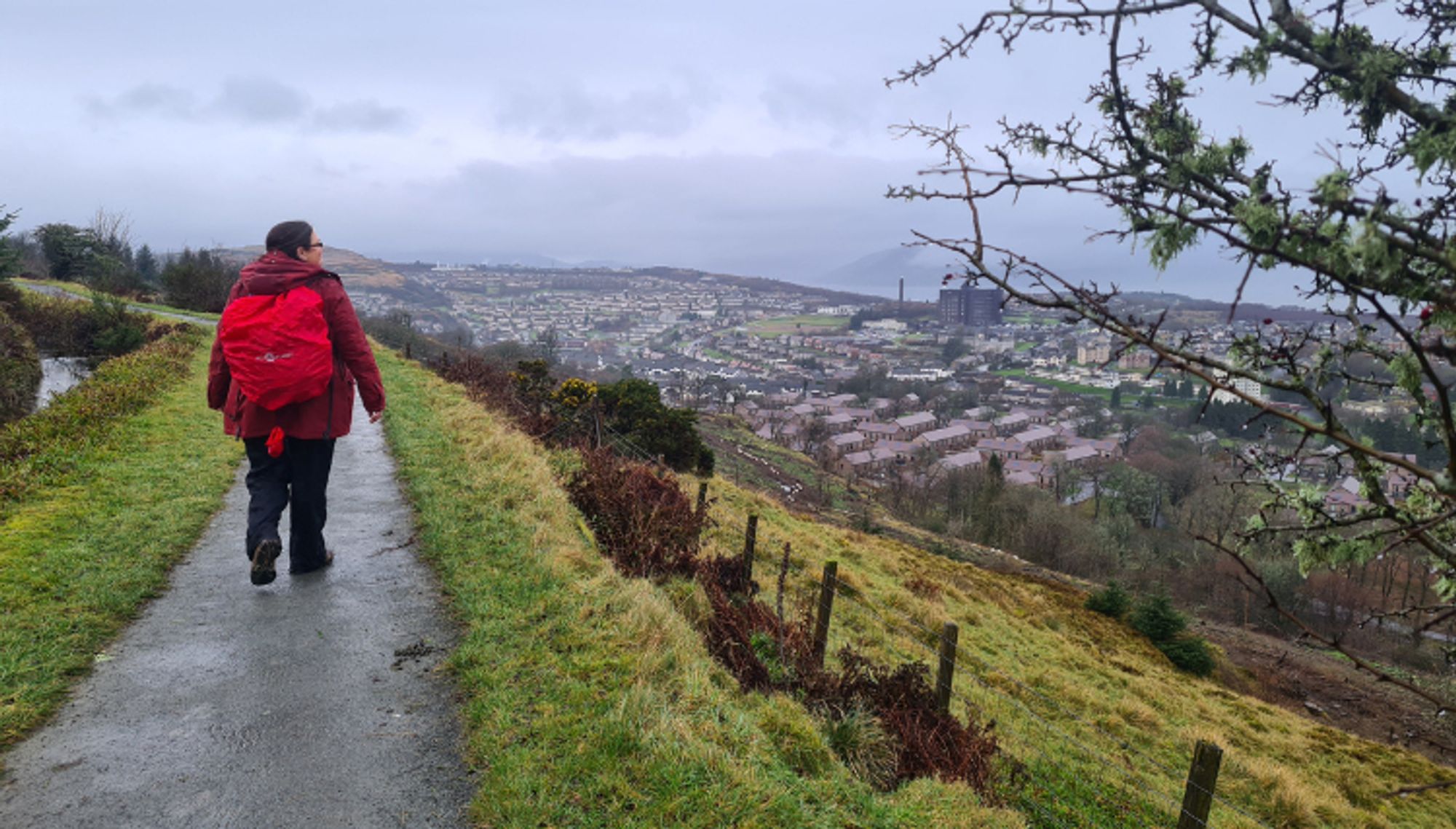 A person wearing a red jacket and backpack is walking along a paved path on a grassy hill, overlooking a town with houses and buildings in the distance under a cloudy sky.