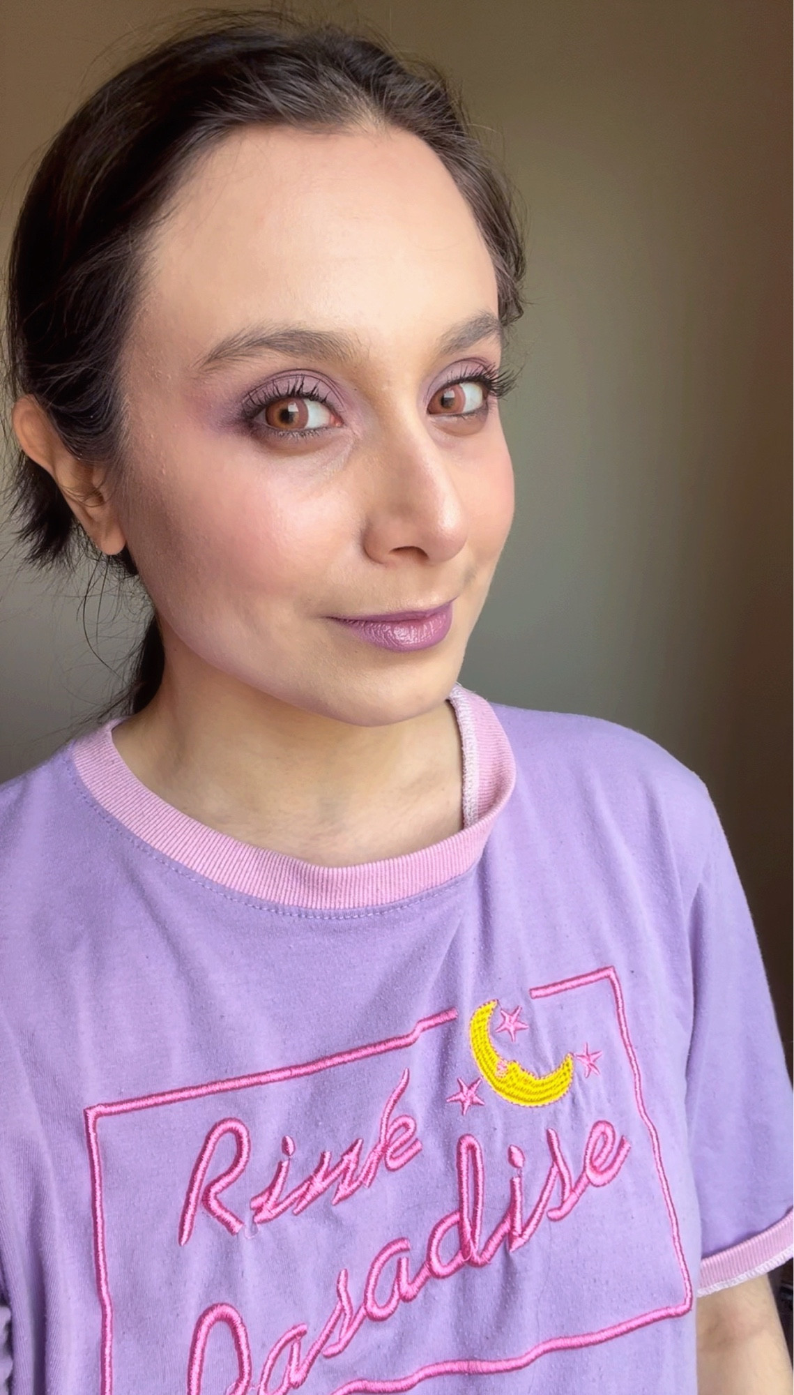 User Juicy Juice is in this image with a pastel lavender and magenta Pink Paradise kpop shirt. He is sporting a light golden lilac plum makeup look, looking certain and gently directly into the camera. His hair is pinned back in a long ponytail.