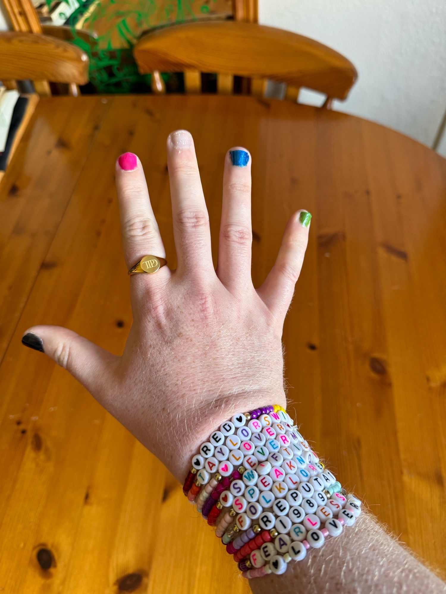 My right hand showing black, pink, silver glittery, metallic navy blue & green nails, gold TTPD cygnet ring and a number of Taylor Swift friendship bracelets