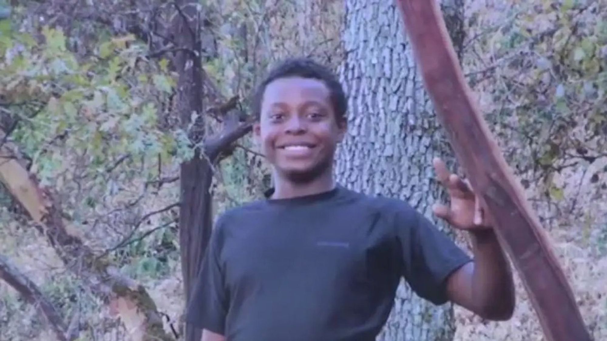 Ryan Gainer, a 15-year-old Black autistic boy wearing a black t-shirt, stands smiling in the woods.