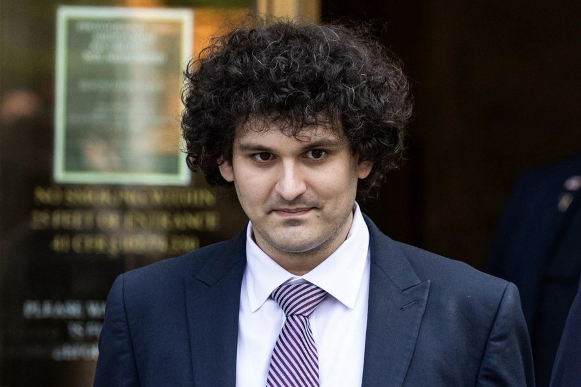 Sam Bankman-Fried, a white man wearing a black suit, white dress shirt and blue-and-white-striped tie, with big scraggly hair, standing outside of a courthouse.