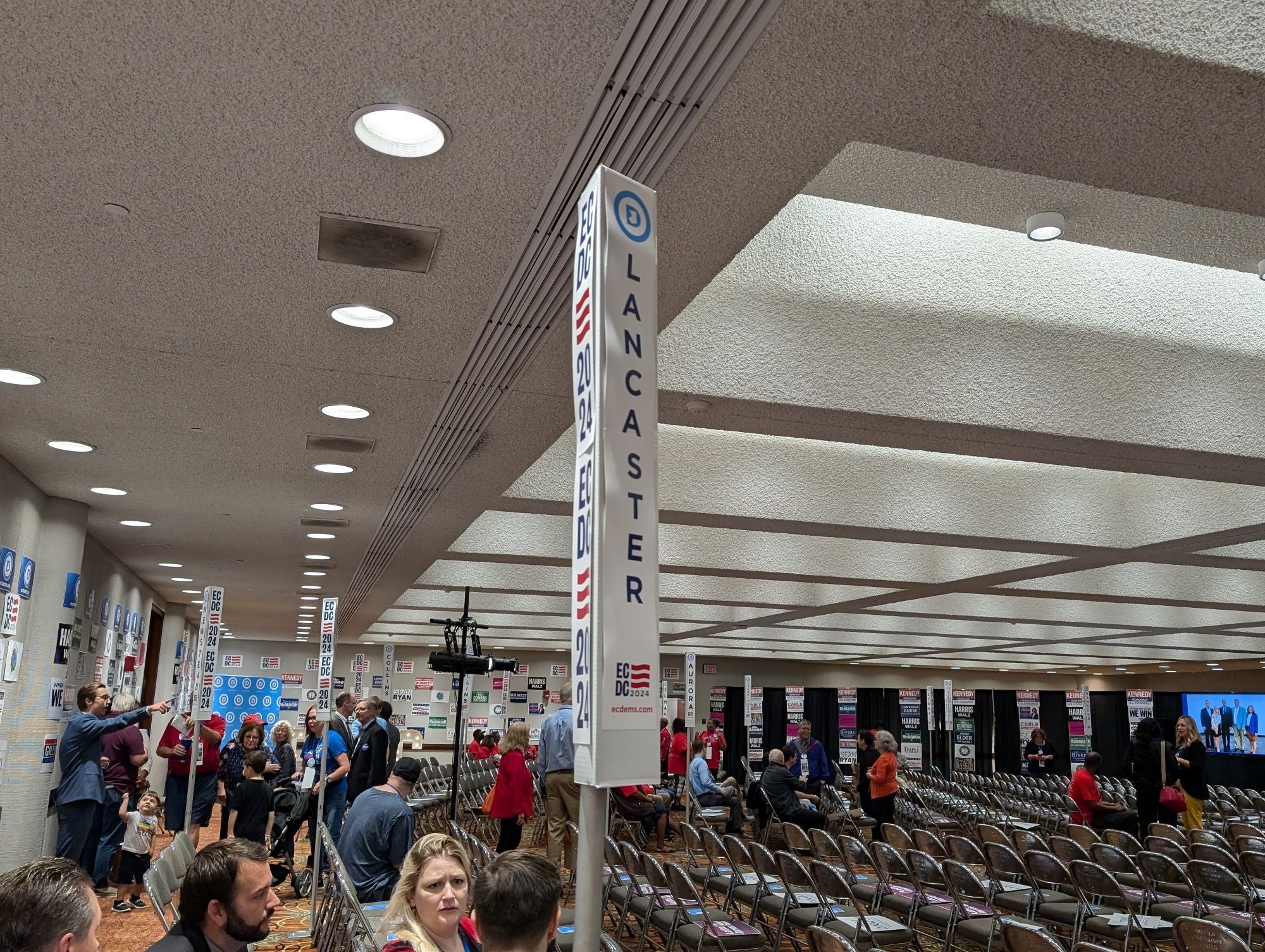 Convention center room filling with people. There are vertical signs like those seen at national conventions with the names of towns.