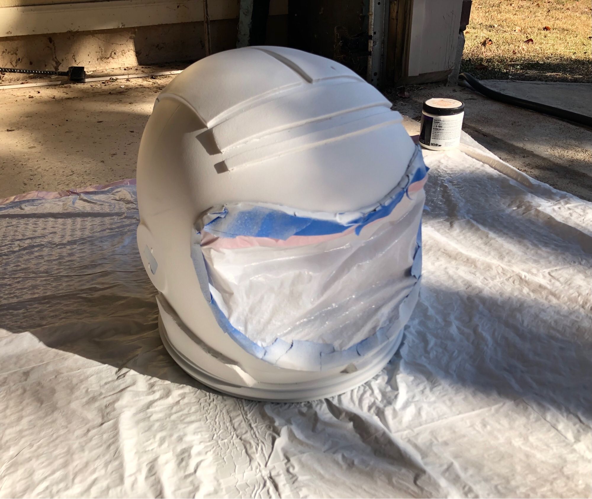 A space helmet sealed with tape and trash bags, covered in white primer and drying in sunlight