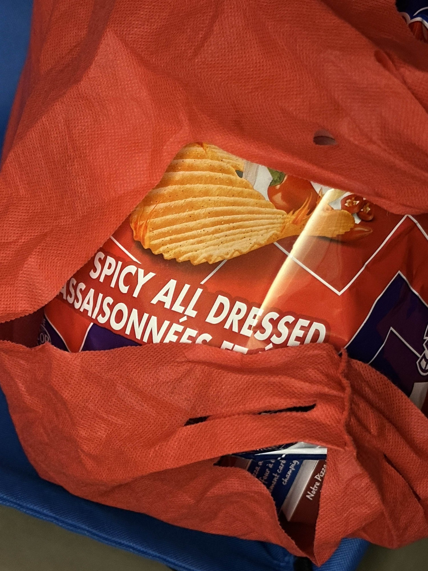 "spicy all dressed" printed in white block lettering runs left to right across the red back ground of a bag of ruffles brand potato chips. above the letters rest a partially skewed picture of a neatly curved chip with flaming edges and beyond it, a red spicy pepper. the rest of the bag is obscured by the handles and folds of a red reusable grocery bag 