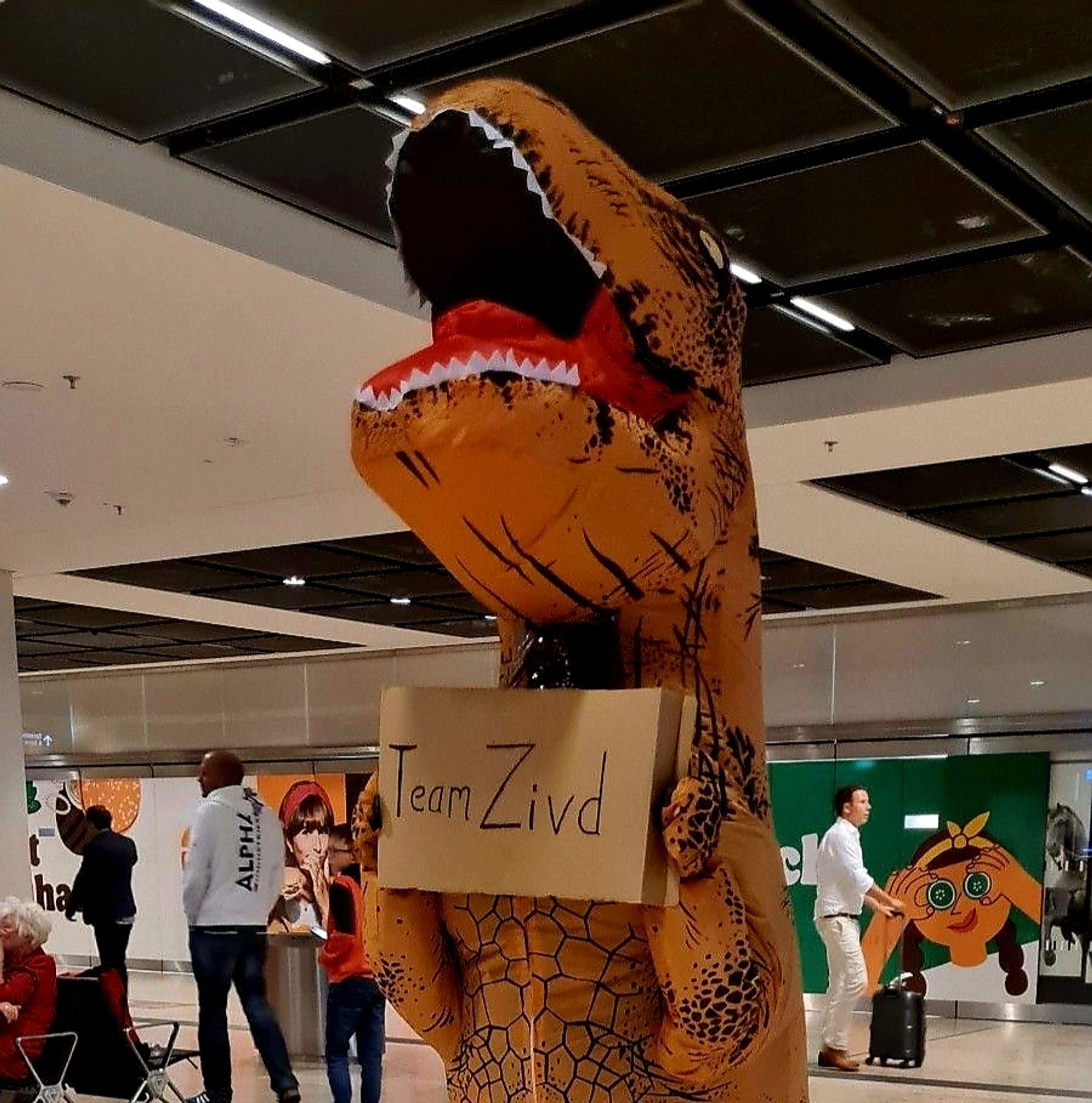 Ich in einem dino kostüm am Flughafen, mit einem Schild in der Hand, drauf steht, Team Z i v d