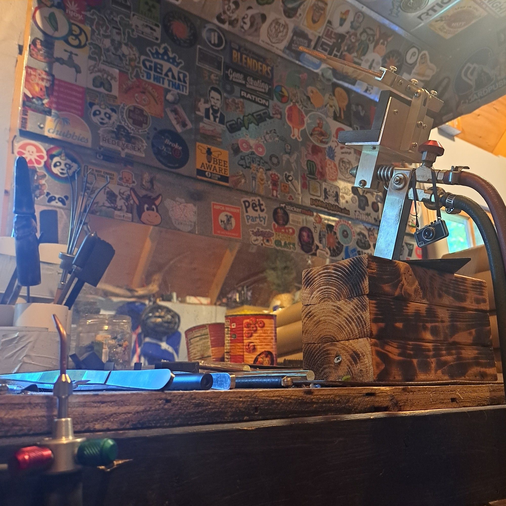 A picture of my work space where I lampwork. It's a wooden bench with some glass working tools on it. A metal vent hood above (with lots of stickers stuck to it) with a big torch mounted to a piece of wood. Also, there is a hand torch hanging from the side of the bench.