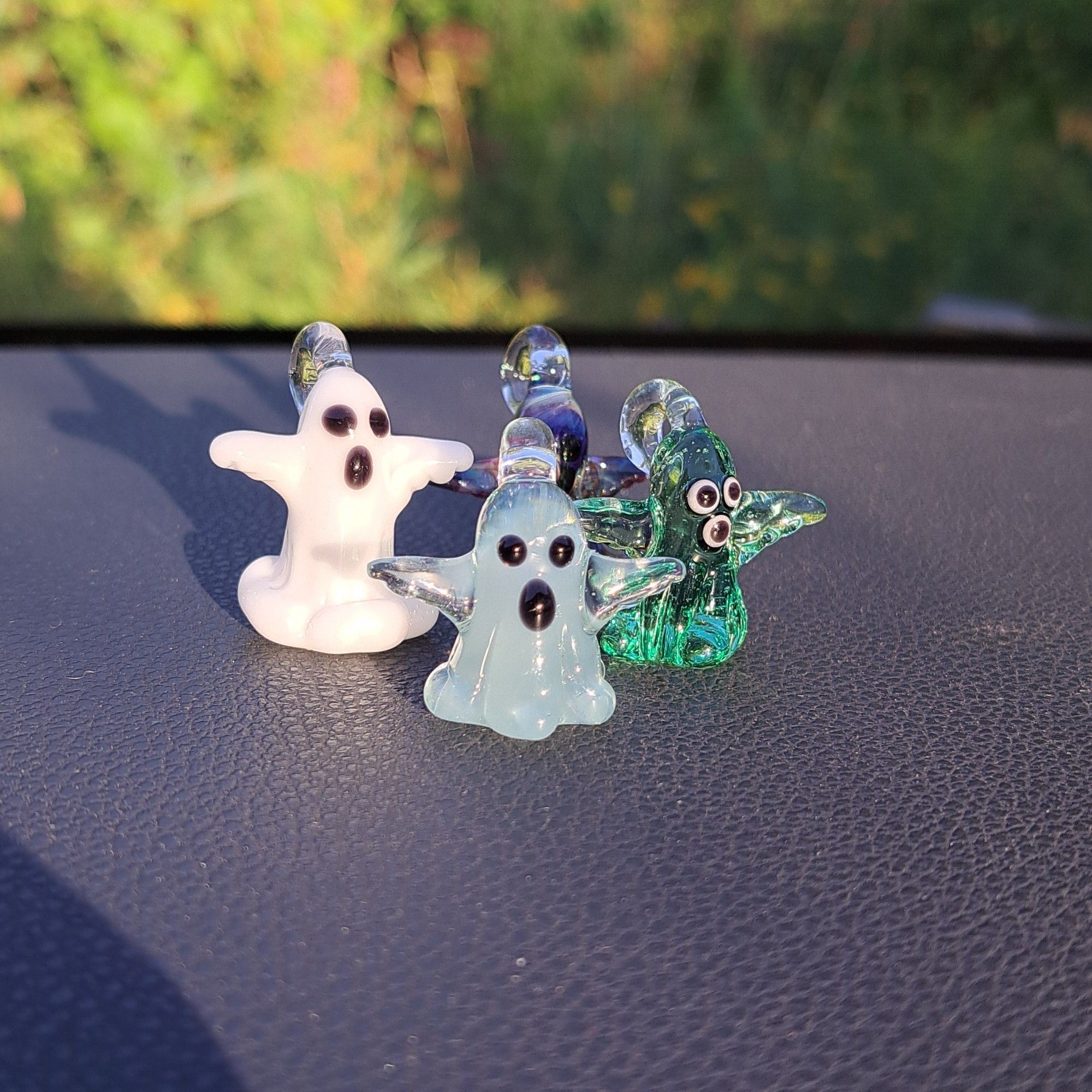 Four little glass ghost pendants sit on a table in a group. All four ghosts are different colors. One is classic white, transparent white, transparent green, and a multicolored one. All ghost have their arms out like, HUG ME!!