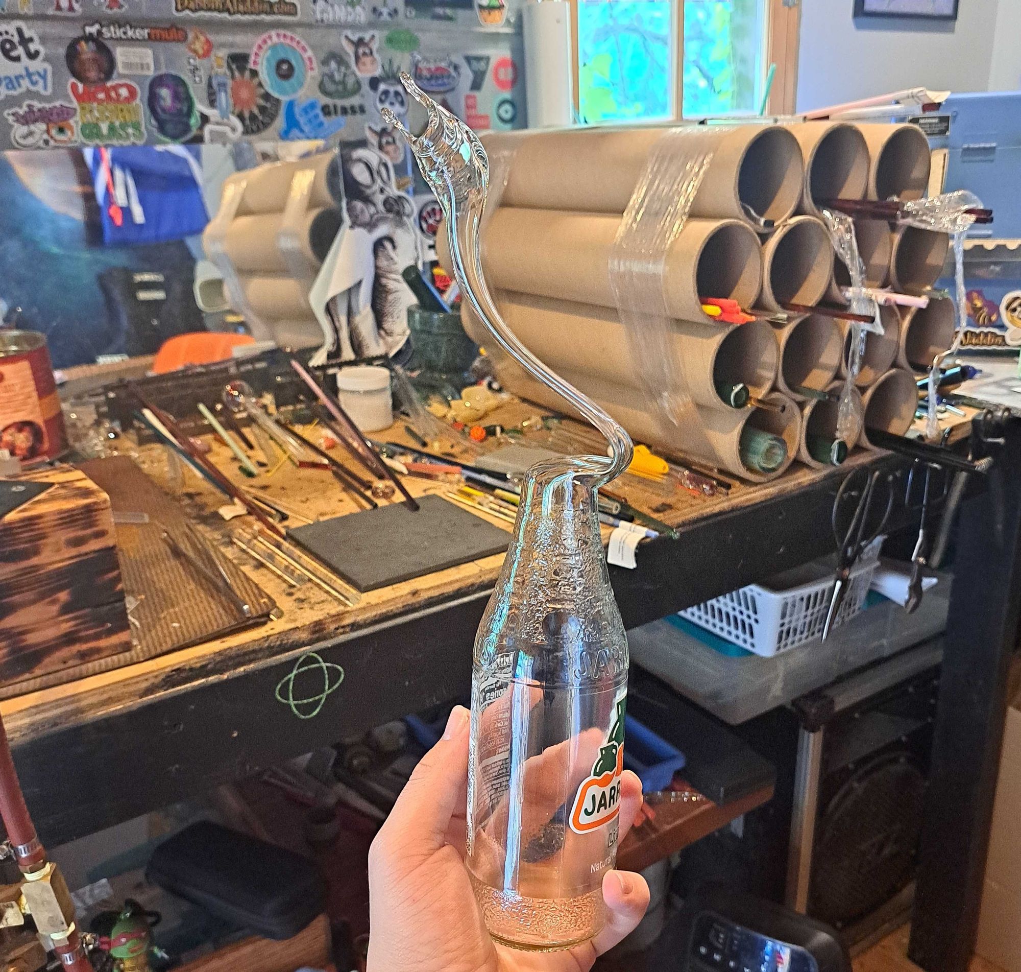 A hand holds an empty glass Jarritos lime soda bottle. The top of the bottle has been manipulated by fire and sculpting to resemble a duck head.  In the background is a messy lampworking bench.